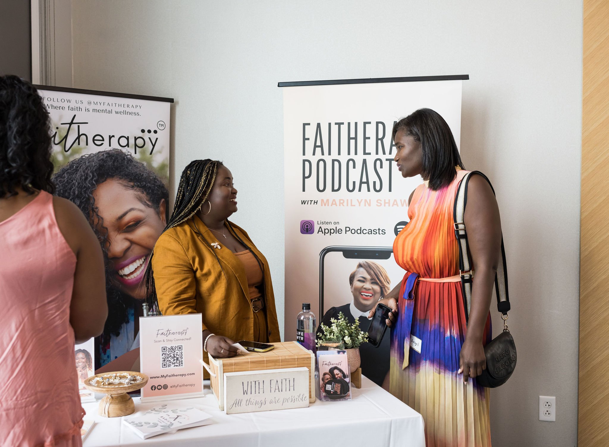 Two women engage in conversation at a podcast promotional booth featuring banners for "Faitherapy Podcast." A table displays promotional materials, and a sign reads, "With Faith, all things are possible." One woman wears a colorful dress, and the oth