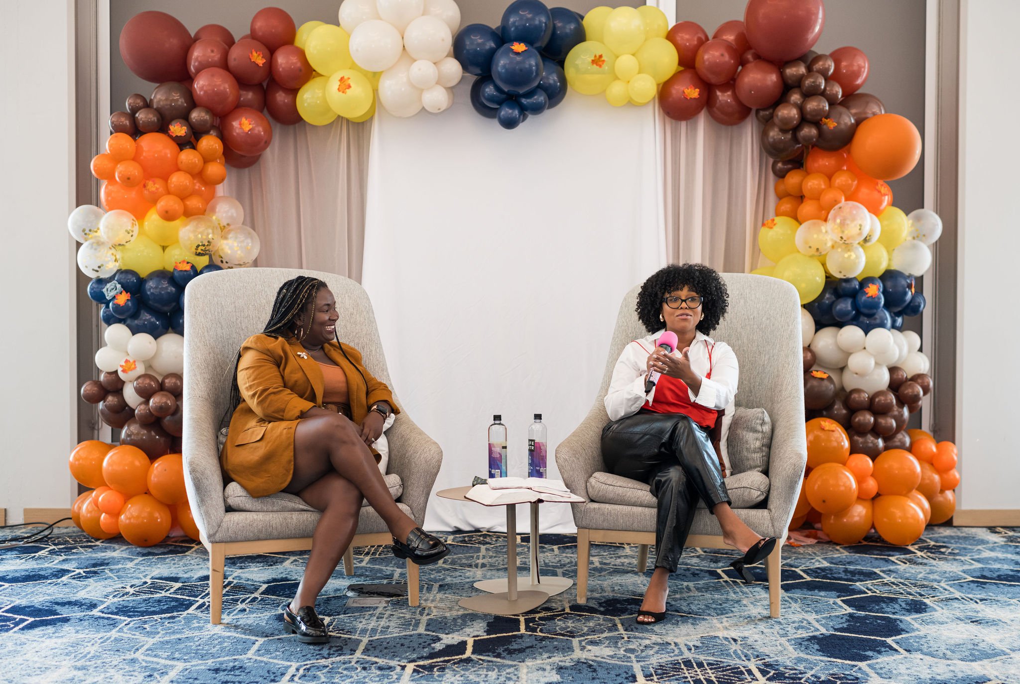 Two women seated in armchairs on a stage, having a conversation. The stage is decorated with colorful balloons in shades of orange, yellow, red, white, blue, and brown.