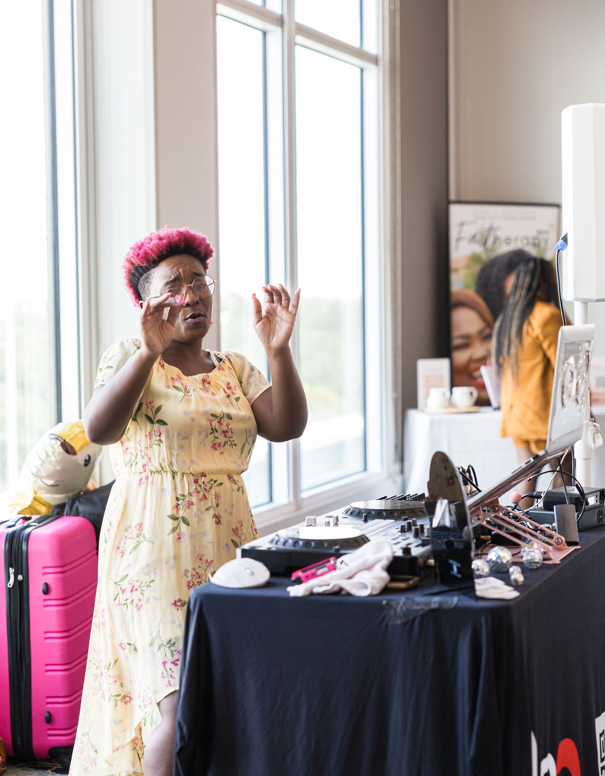 A person with pink hair stands near a table with DJ equipment. They are wearing glasses and a yellow floral dress, with a pink suitcase visible nearby. The room has large windows and a poster in the background.