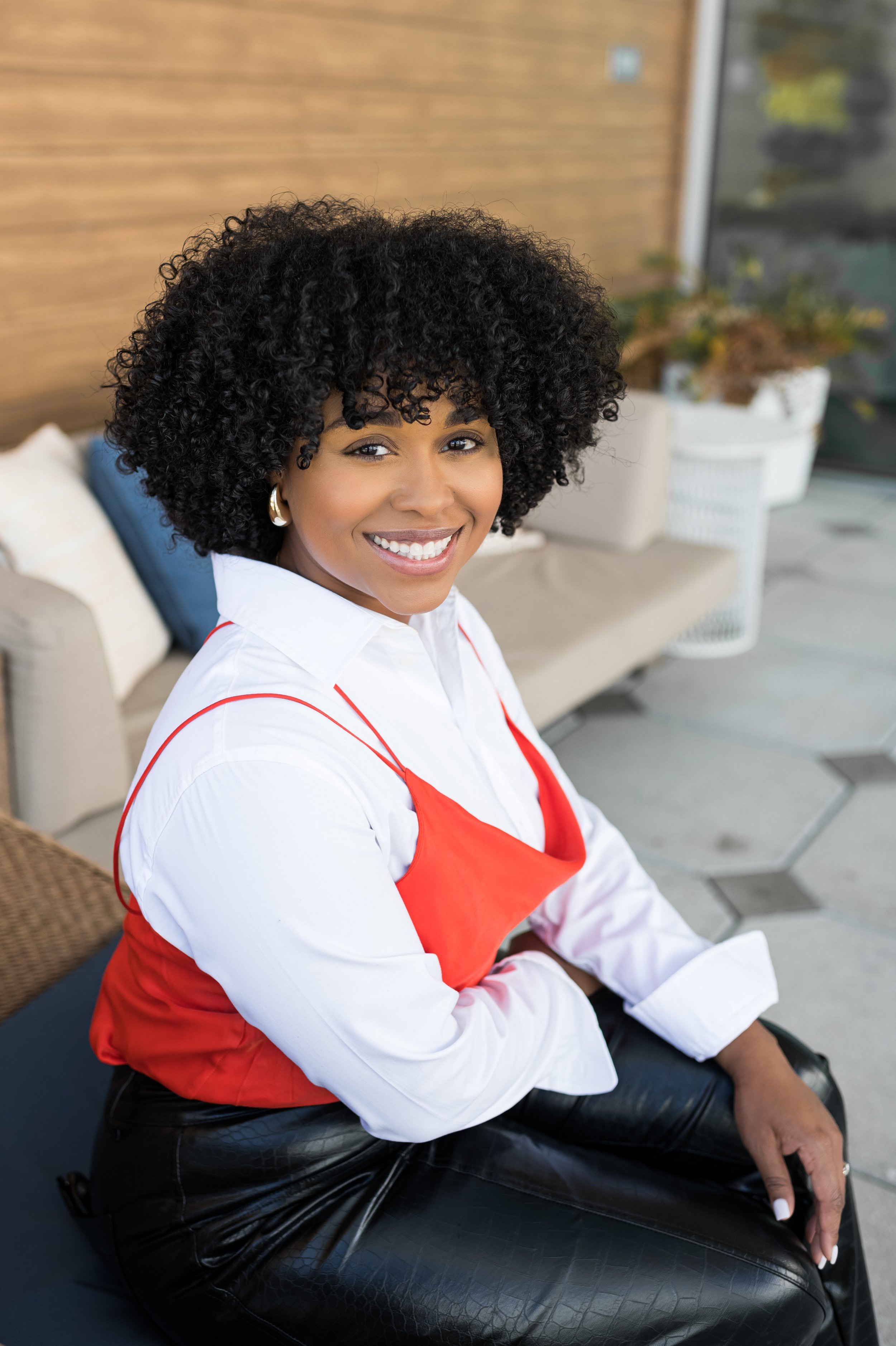 Smiling person with curly hair wearing a red top over a white shirt and sitting outdoors.