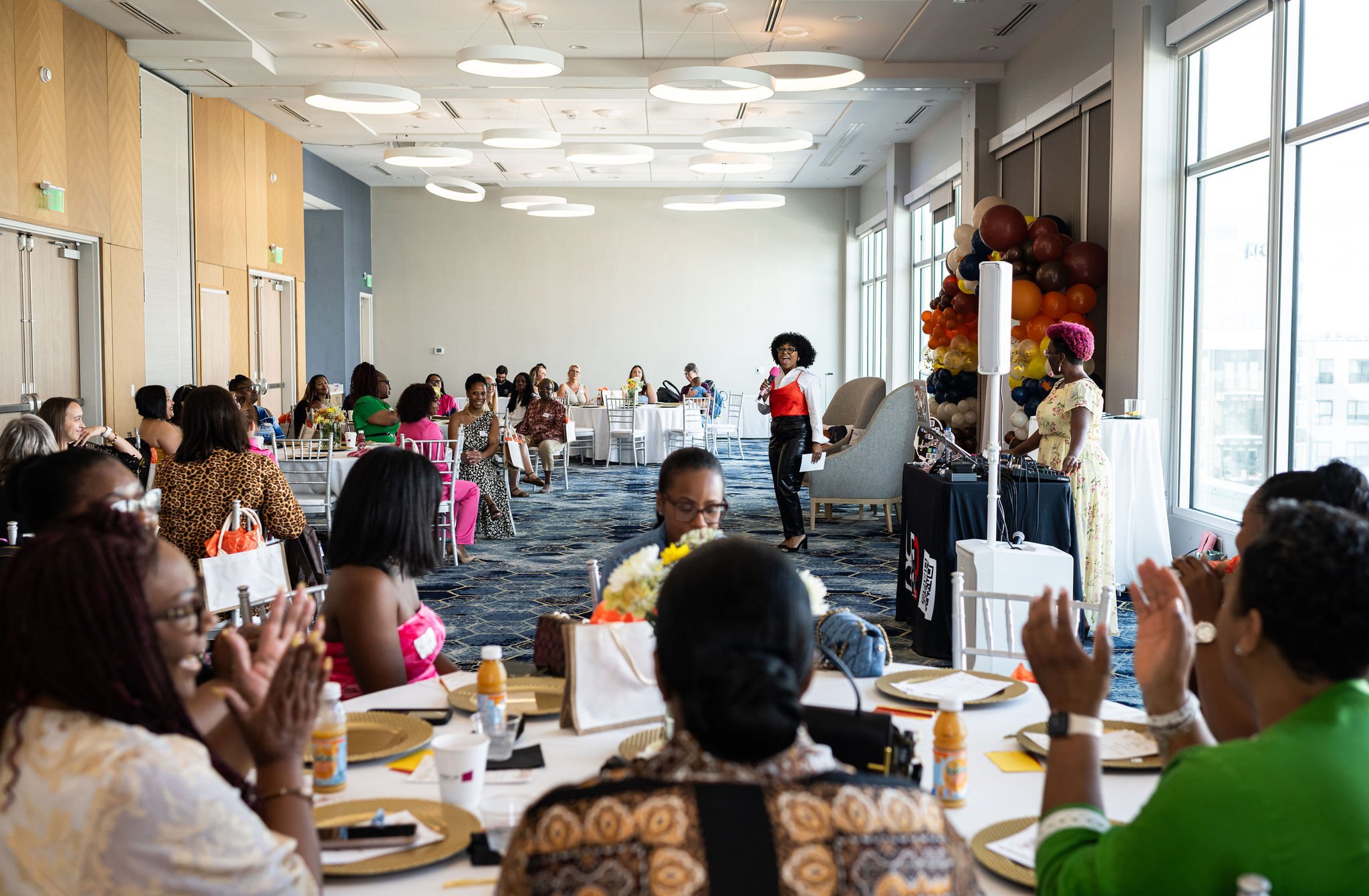 A diverse group of people sitting at round tables in a well-lit conference room, listening to a speaker in front. The room is decorated with a balloon arrangement, and attendees are engaged, with some clapping. The atmosphere appears formal and busin