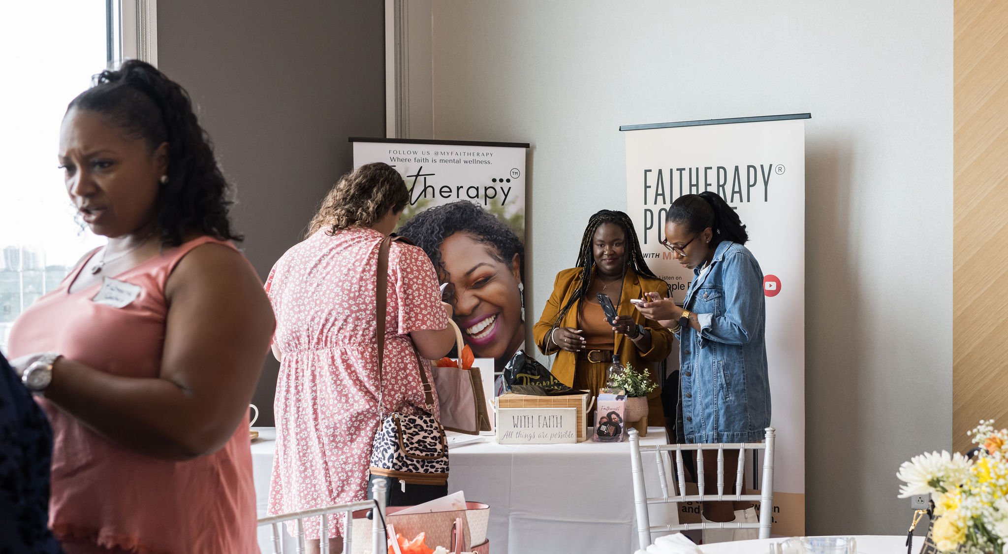 Women interacting at a booth with 'Faith Therapy' and 'Faith Therapy Podcast' banners, featuring a vase of flowers on a nearby table.