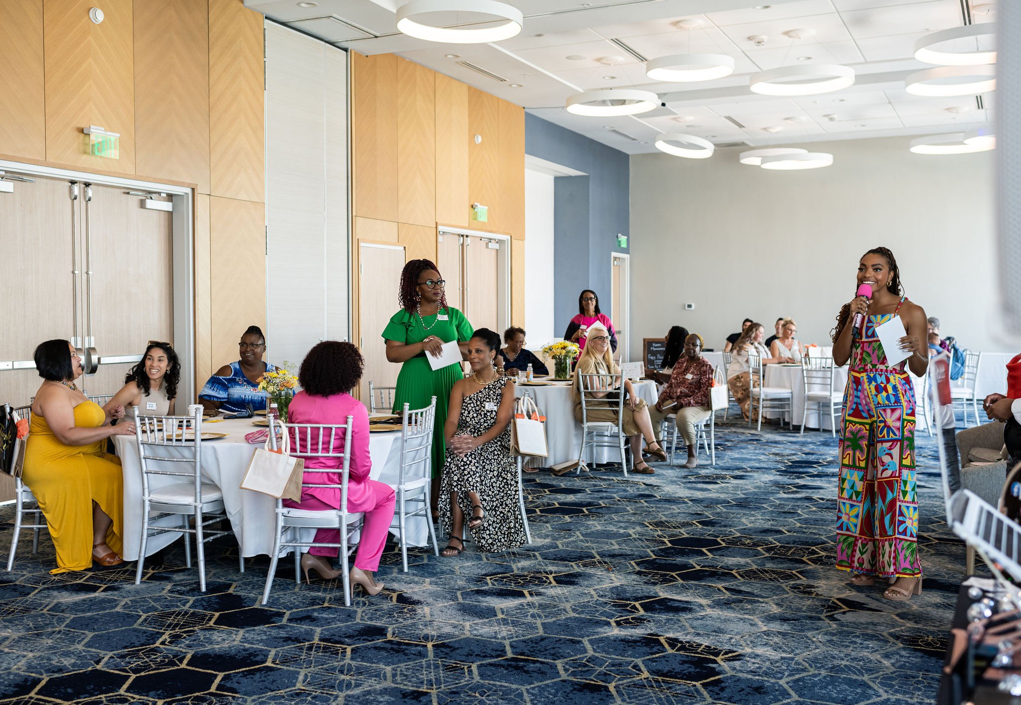 A group of people gathered in a modern conference room, sitting at white-clothed tables with white chairs. A woman in colorful attire is speaking into a microphone, holding a note. The room has a blue carpet with a geometric pattern and overhead circ