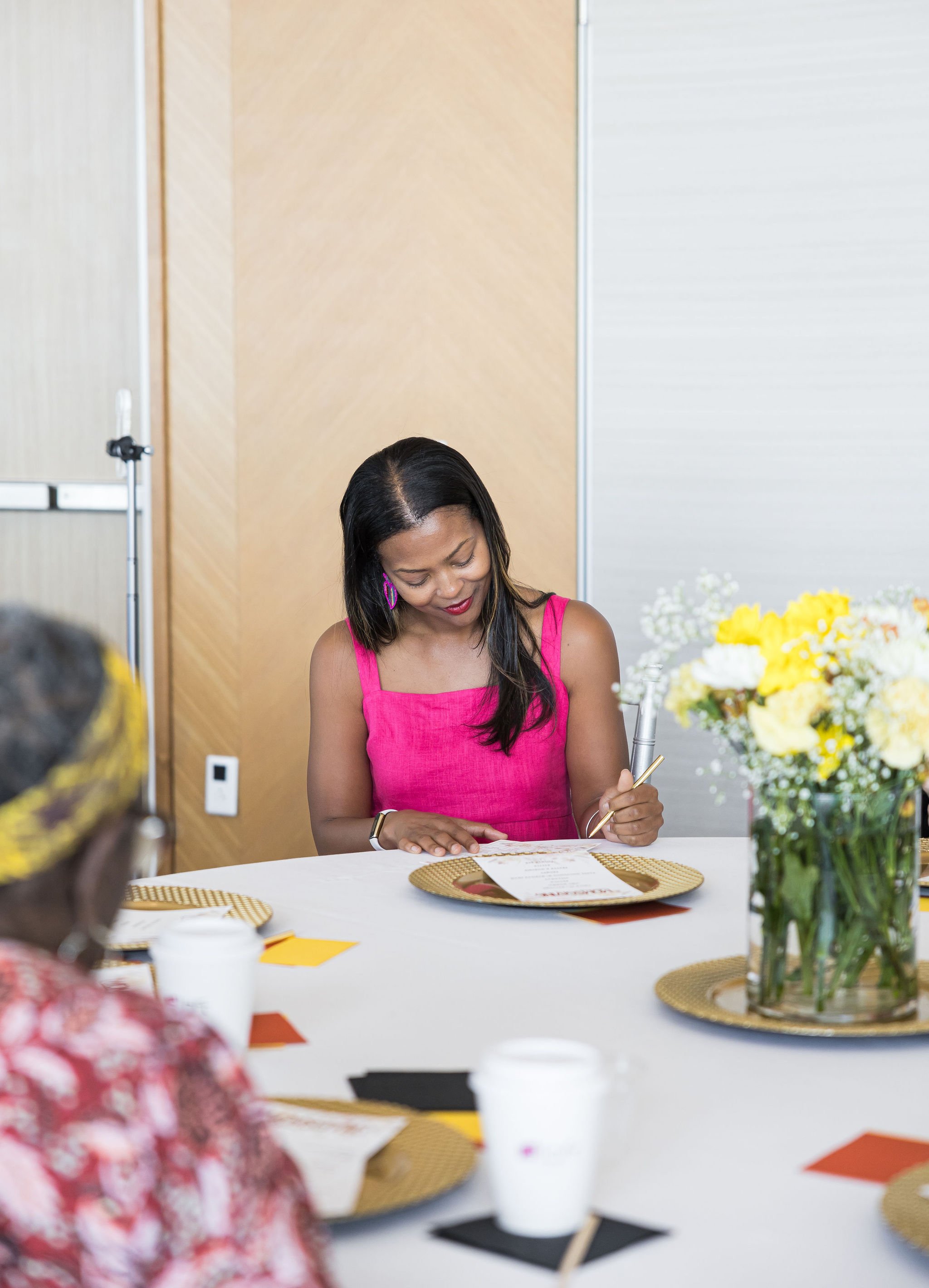 Woman in pink dress writing at a table with flowers and papers