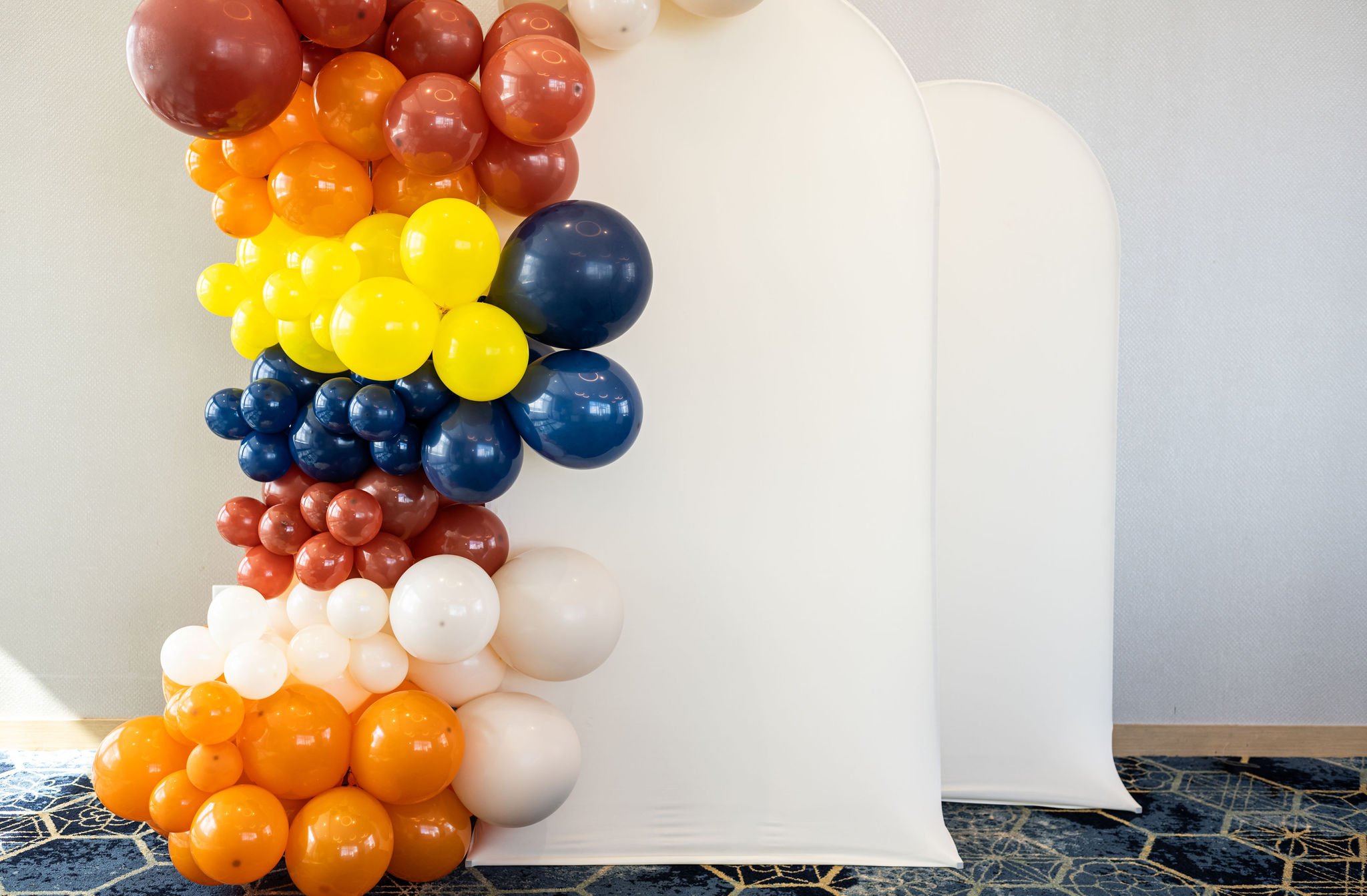 Colorful balloon arch next to a white backdrop in a room with patterned carpet.