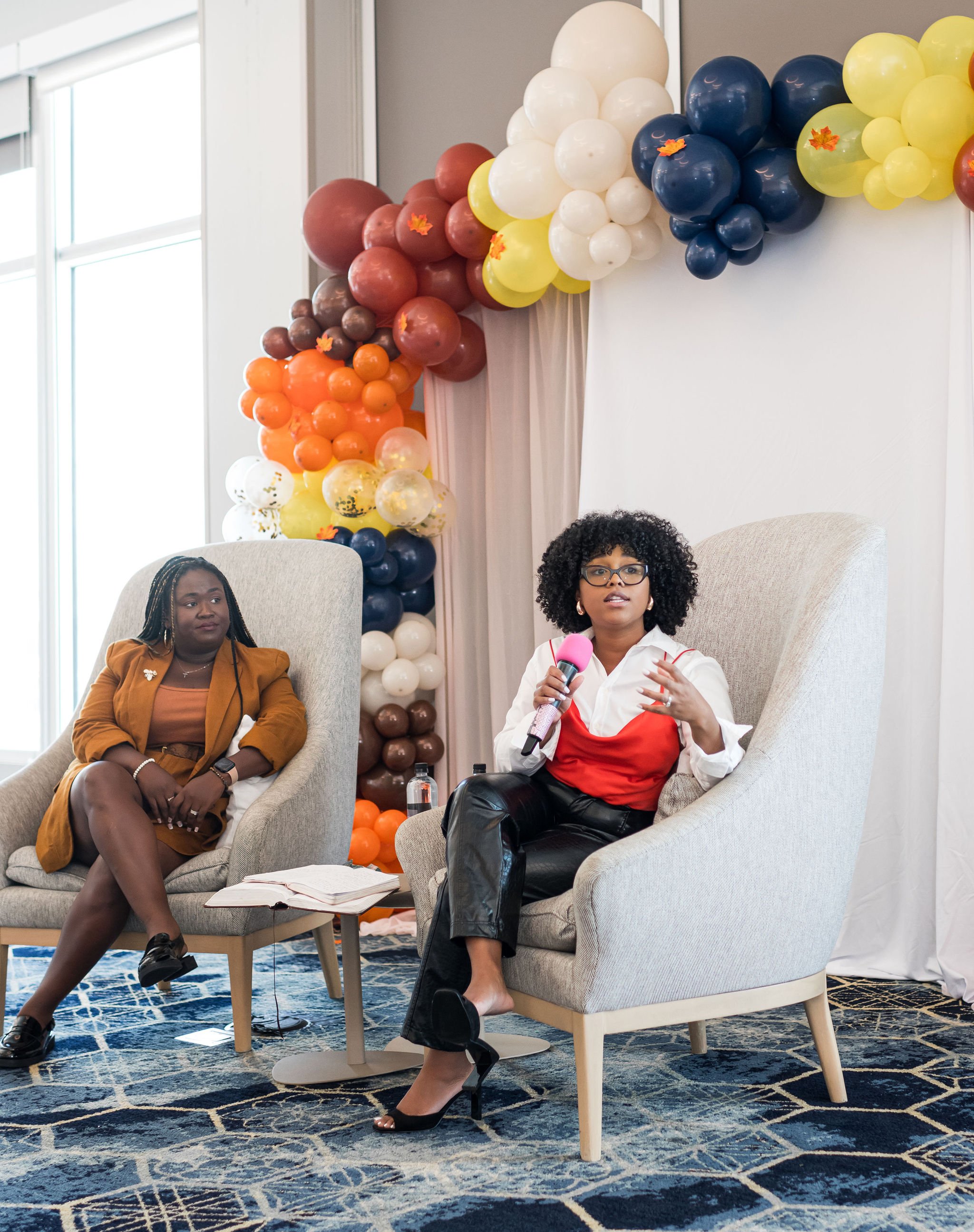 Two women seated in armchairs with colorful balloon decorations in the background, one holding a microphone, engaged in a discussion.