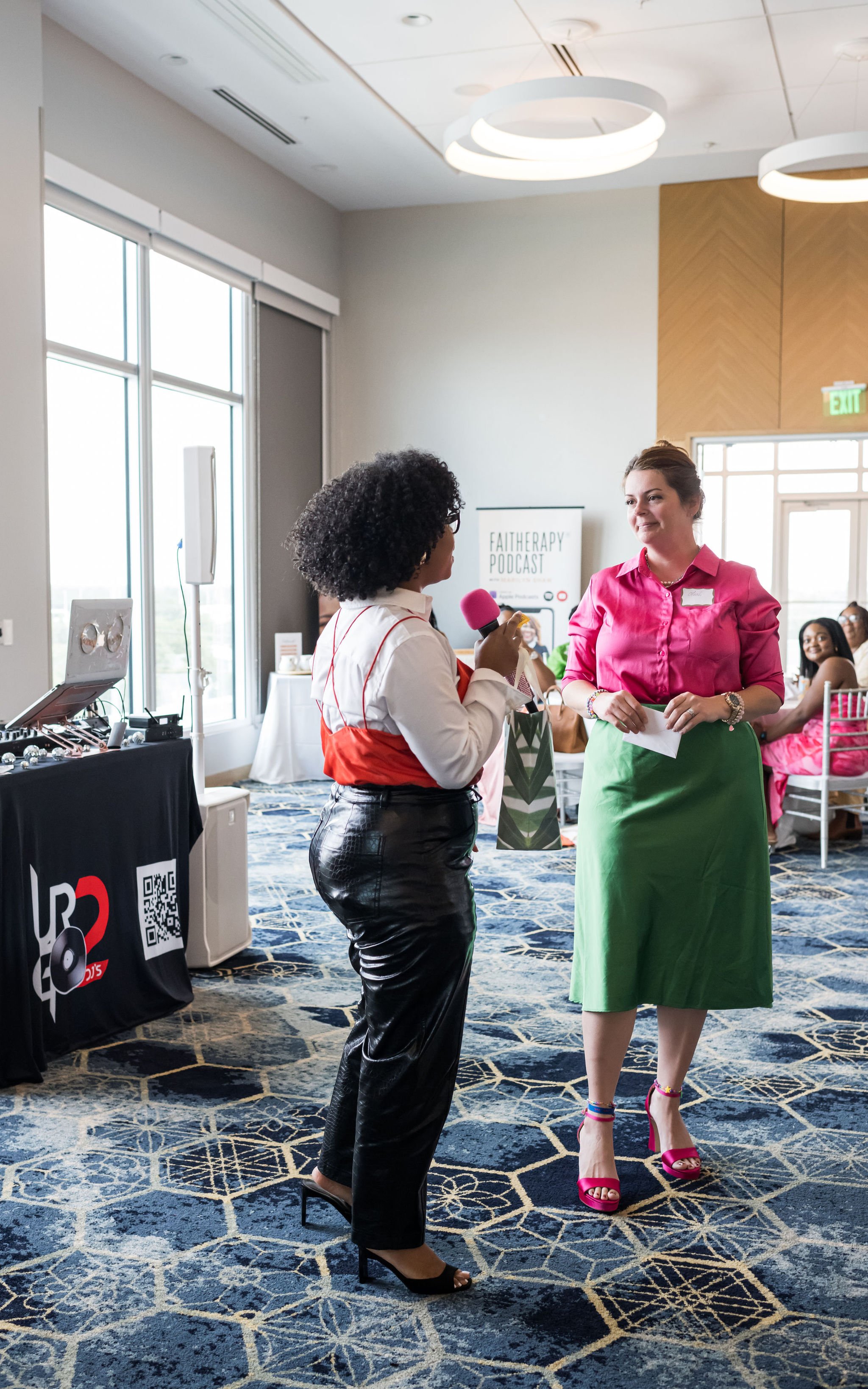 Two women in a meeting room, with one holding a pink microphone, standing near a table with a laptop and speakers. A "FaithErapy Podcast" banner is visible in the background.