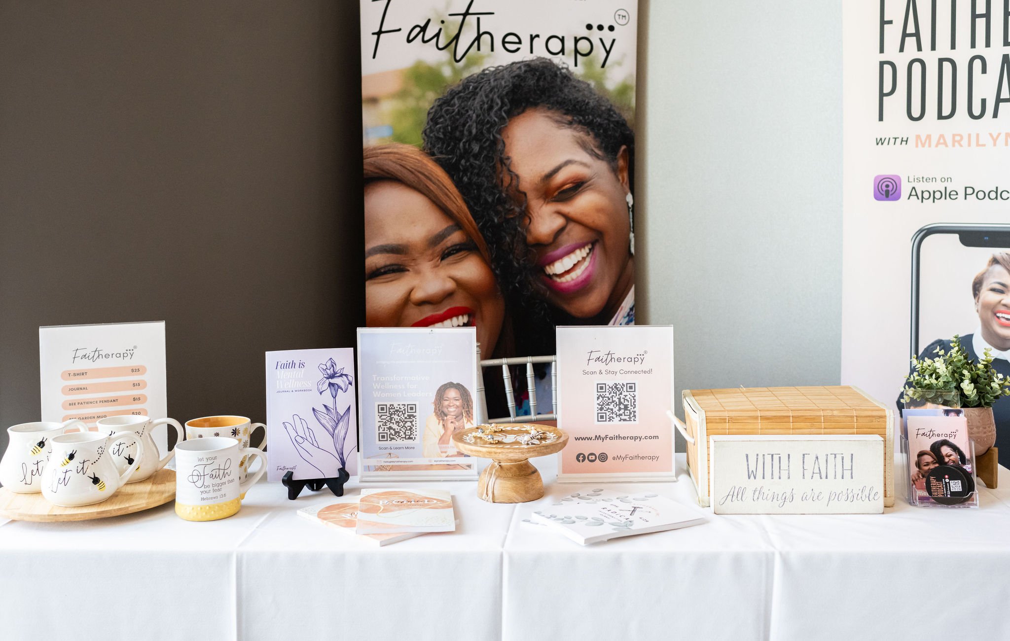 Display table featuring "Faith Therapy" promotional materials, mugs, brochures, and a sign with a motivational quote. Includes price list, QR codes, a wooden dish with jewelry, and a podcast advertisement. Background with a large poster of smiling wo
