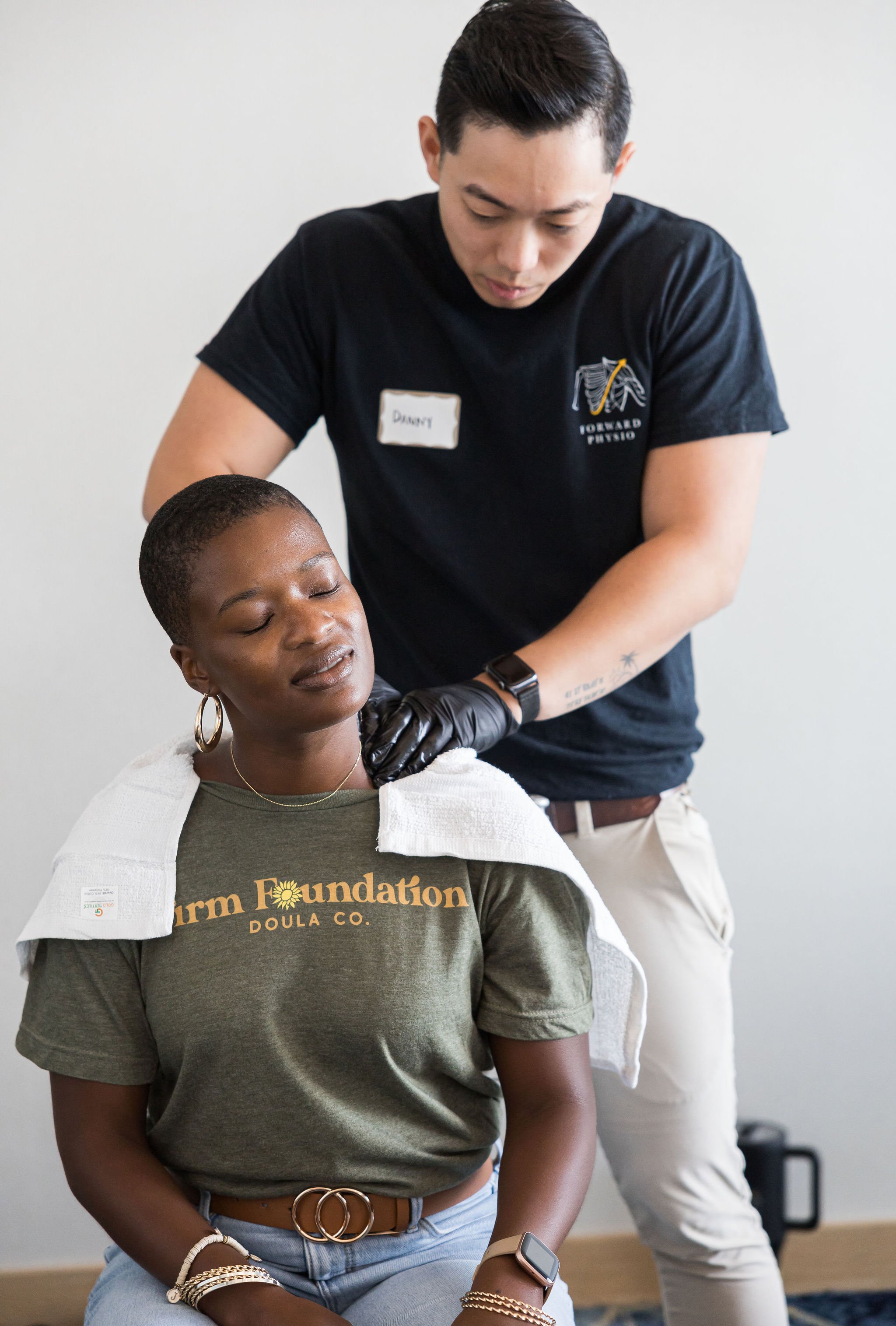 A man wearing gloves is giving a neck massage to a woman seated with her eyes closed, wearing a t-shirt with "Farm Foundation Doula Co." written on it.