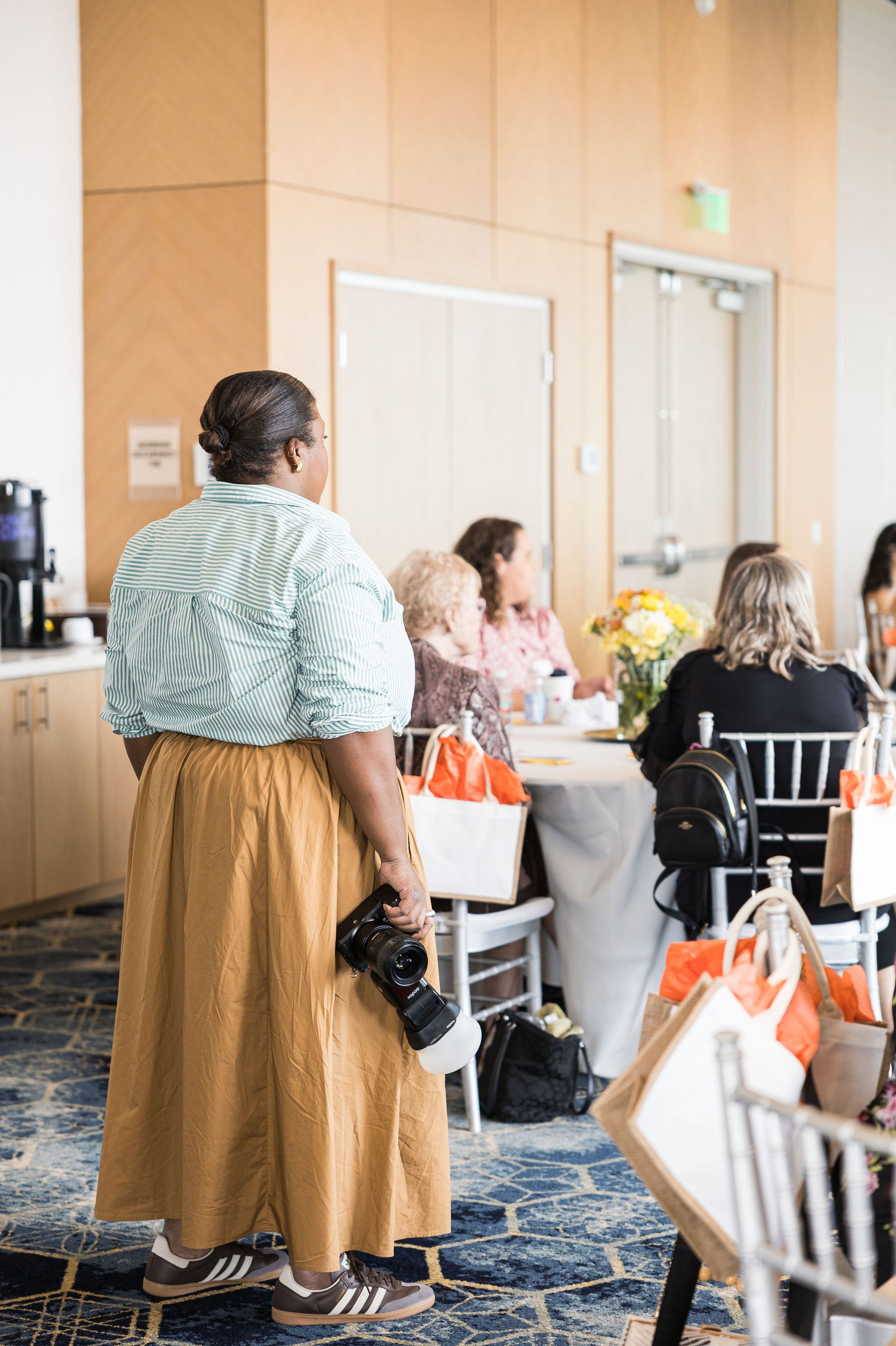 Person holding a camera in an event room, with people seated at tables, floral arrangements, and gift bags visible.