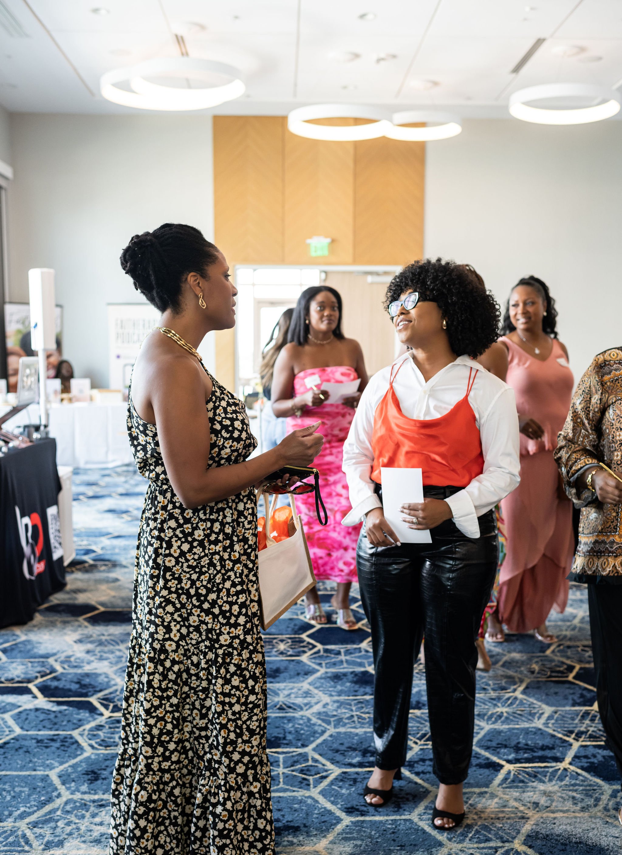 Group of women in a stylish indoor event, with modern lighting and carpet, engaging in conversation.