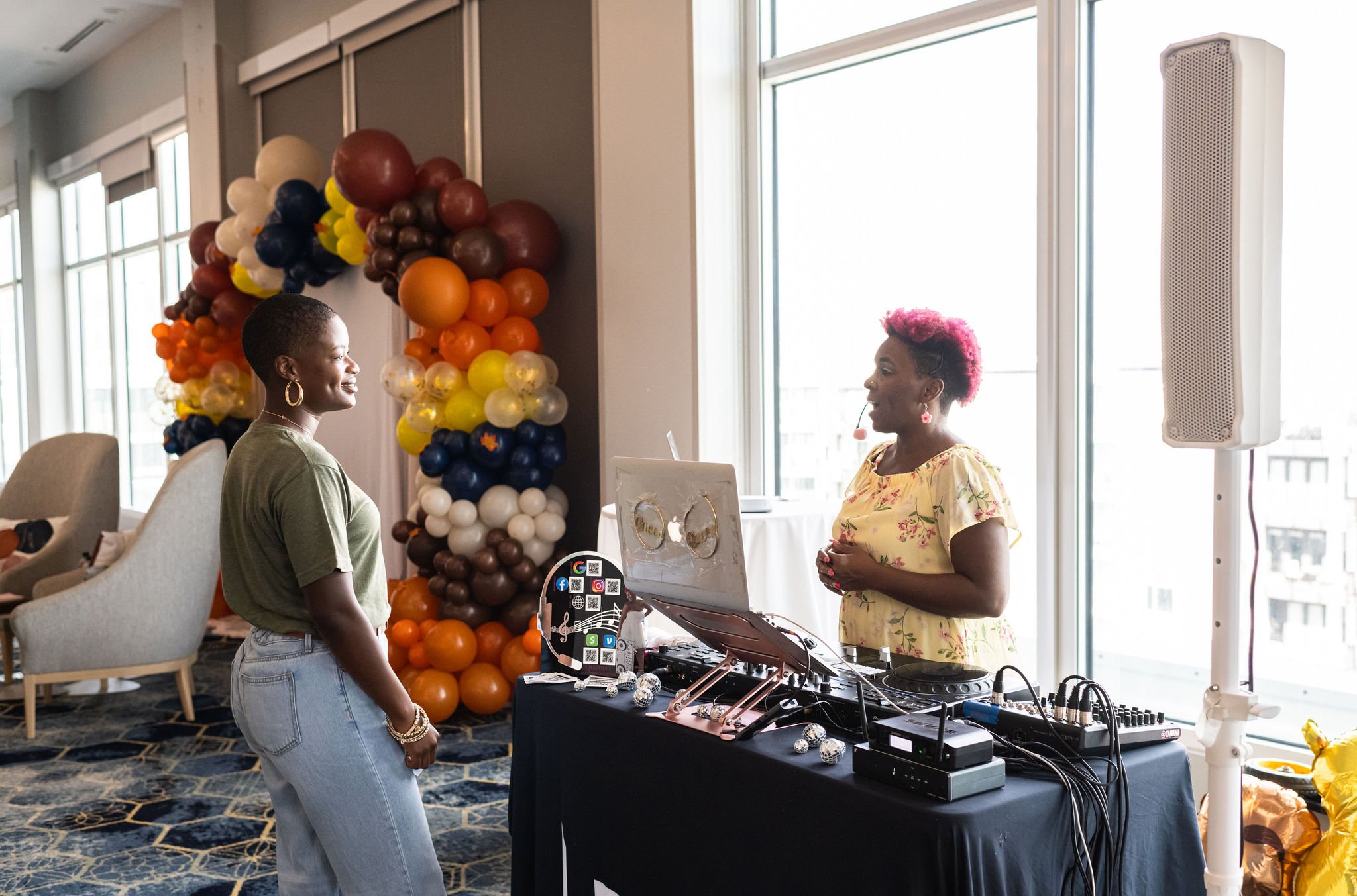 Two women interacting at an event; one is behind a DJ booth, with colorful balloons in the background.