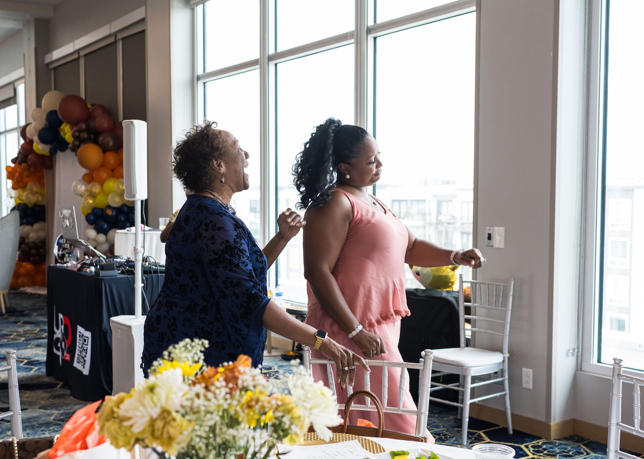 Two women engaging in conversation by large windows in a decorated event space, with floral arrangements and balloon decorations in the background.
