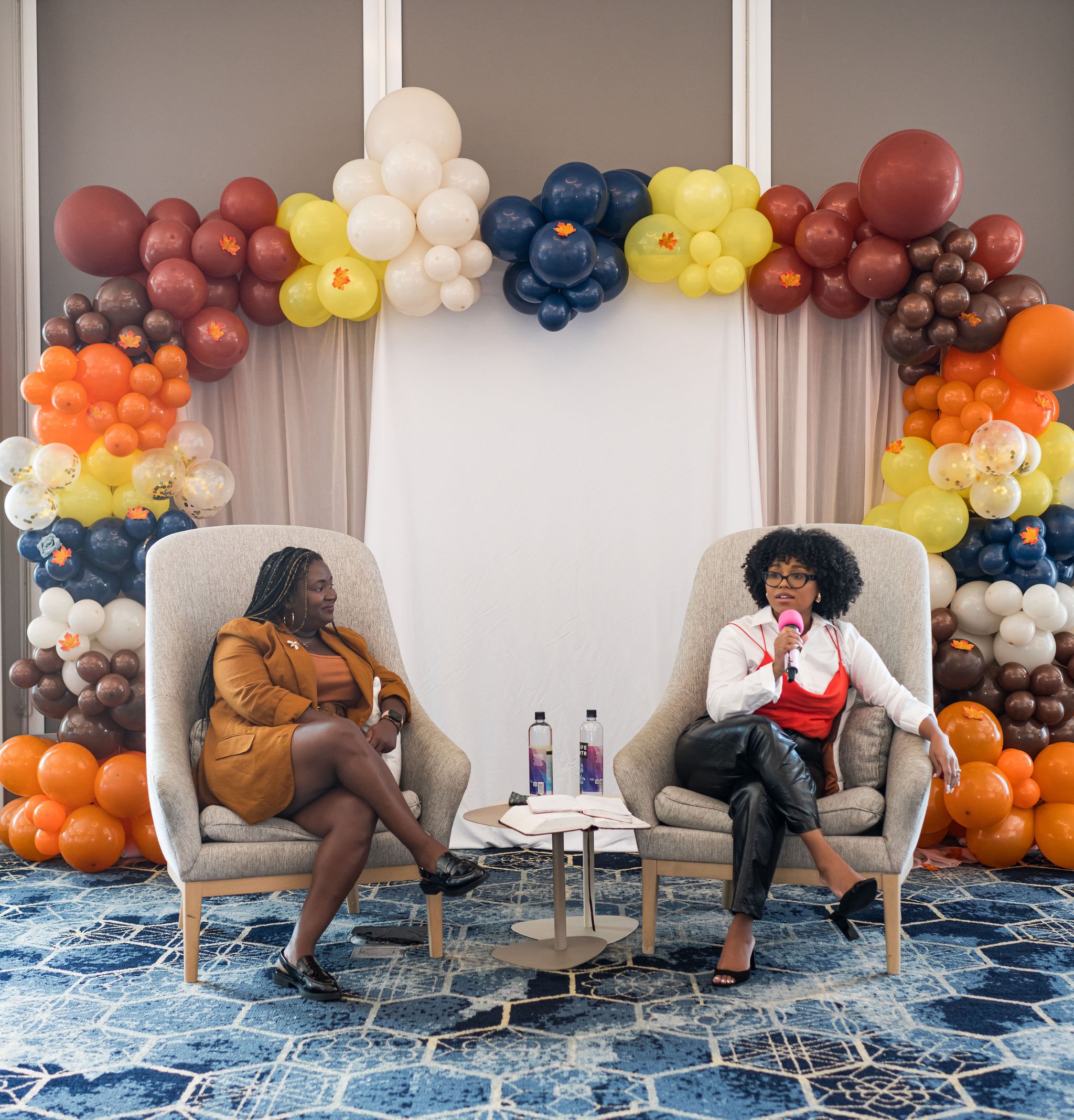 Two women sitting in chairs under a colorful balloon arch, one with a microphone, on a blue patterned carpet at an event.