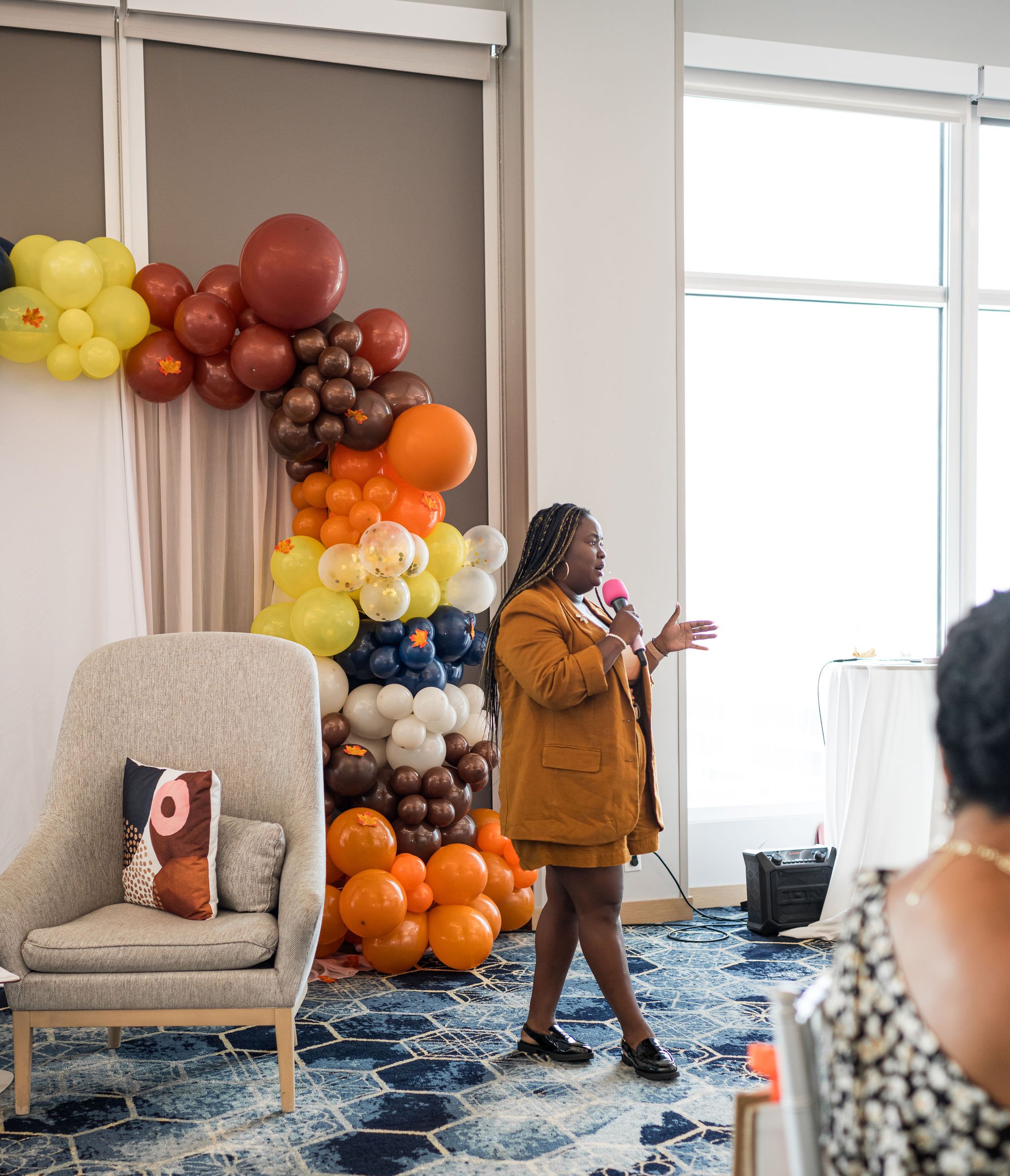 Person speaking with a microphone in a decorated room with a colorful balloon arch and a beige armchair with a patterned pillow.