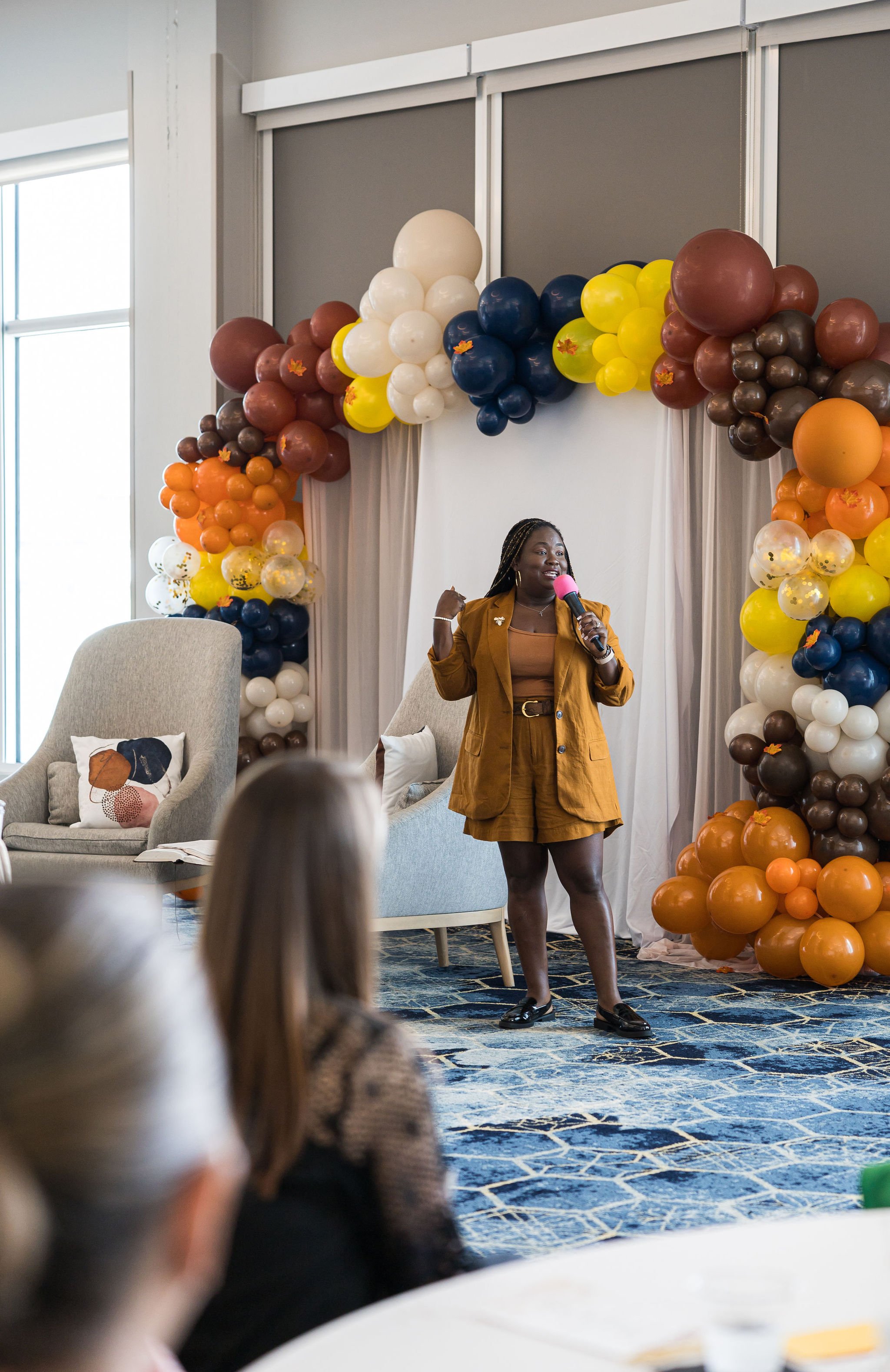 A person speaking on stage with a microphone in front of a balloon arch in autumn colors, including orange, yellow, and brown. The backdrop is set in a room with large windows and an audience is partially visible in the foreground.