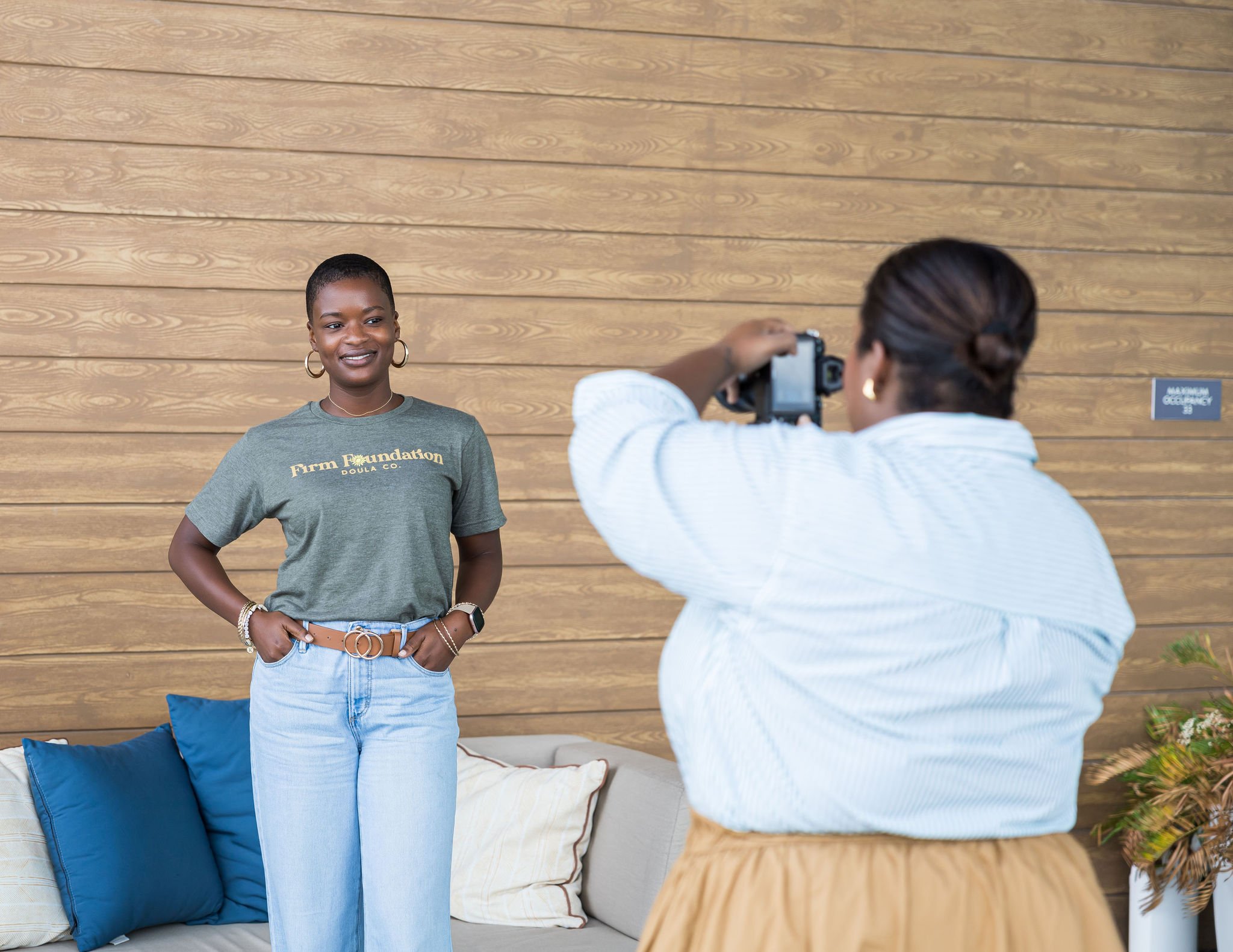 A woman poses for a photo, wearing a green "Firm Foundation Doula Co." t-shirt and light blue jeans, standing in front of a wooden wall. Another person is taking her picture with a camera. A sofa with blue and beige cushions is on the left.