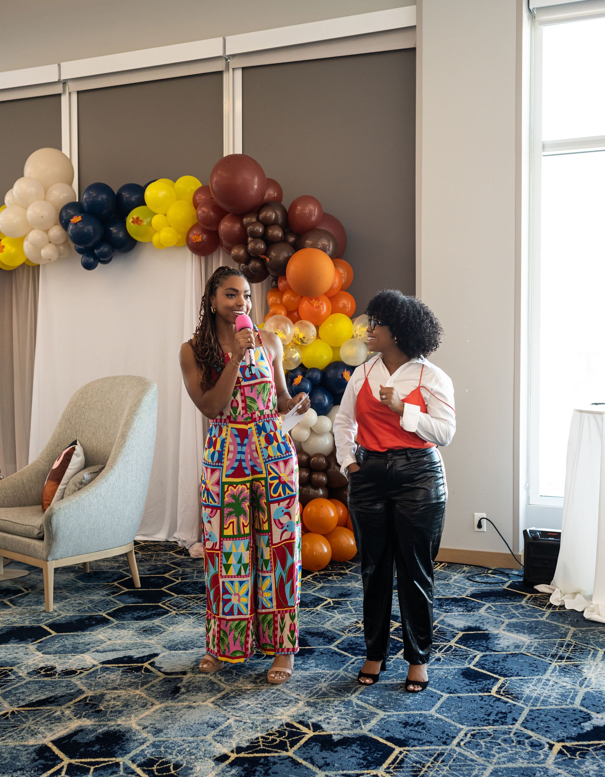 Two women standing indoors near a colorful balloon arch. One holds a microphone and wears a brightly patterned jumpsuit, while the other wears a white shirt with a red top and black pants. A chair is visible in the background on a patterned carpeted
