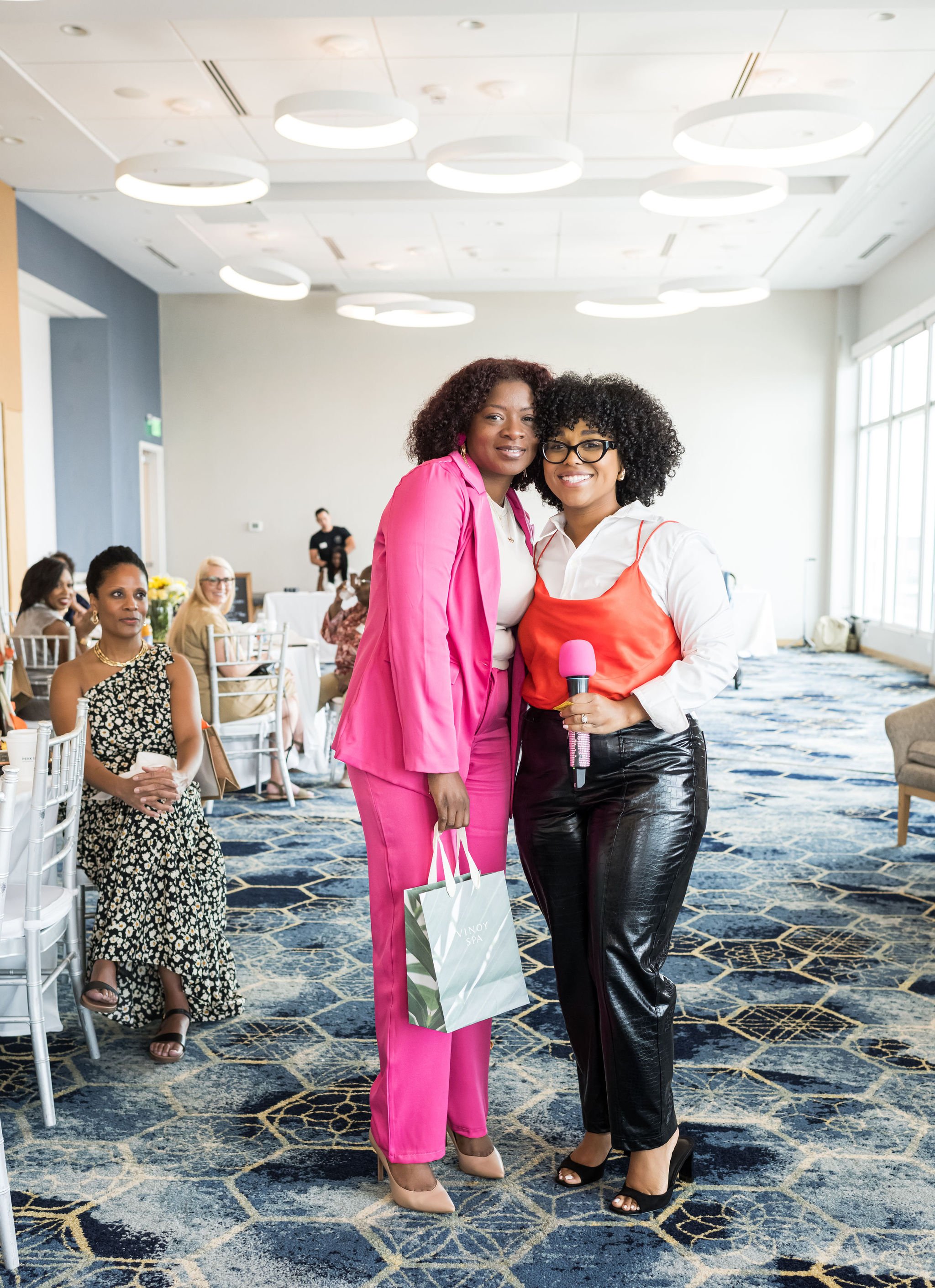 Two women posing together in a brightly lit room with white chairs and people in the background. One woman is wearing a bright pink suit and holding a gift bag, while the other is in a white shirt with a red top and black pants, holding a microphone.