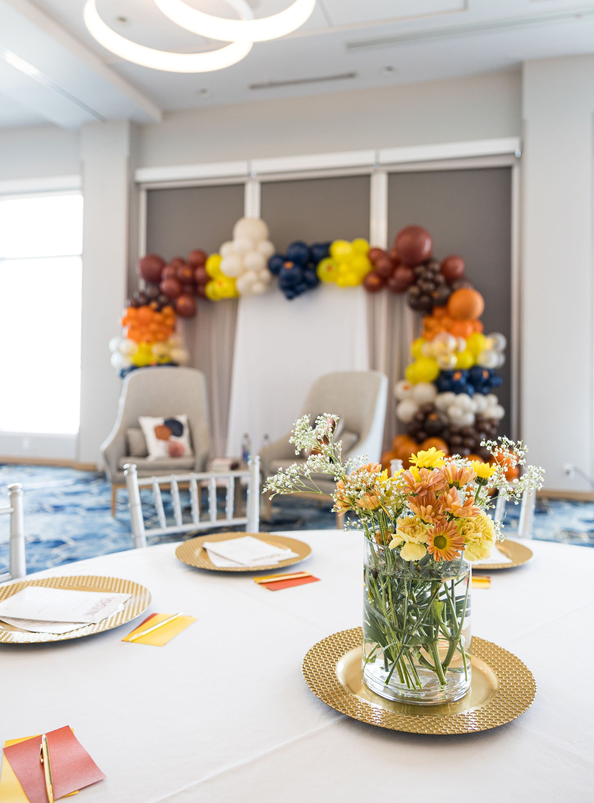 Room decorated with a colorful balloon arch, two chairs, and a table with flowers and gold chargers.