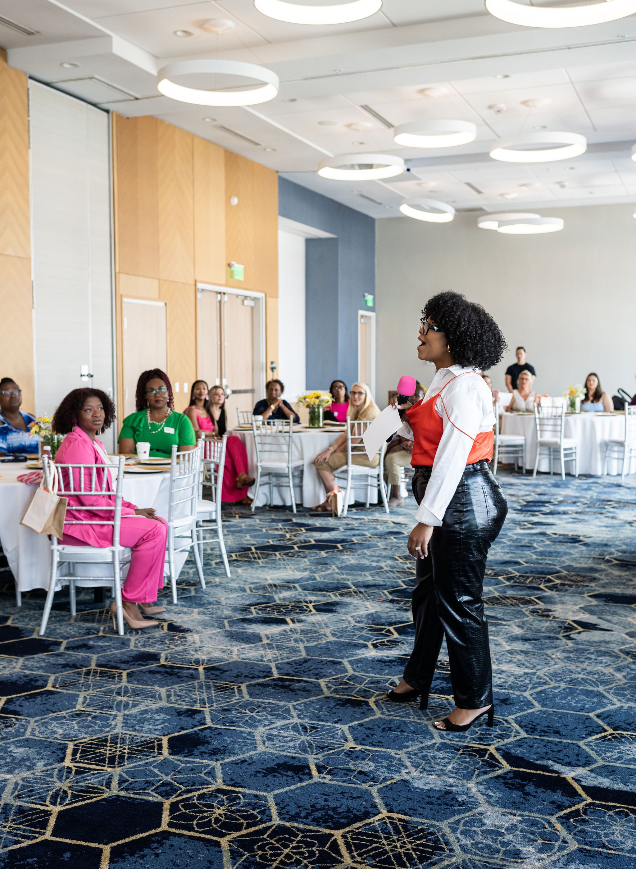 A person is speaking to a group of people seated at tables in a conference room. The attendees are sitting on white chairs around tables with white tablecloths and yellow floral centerpieces. The speaker is holding a pink microphone. The room has mod