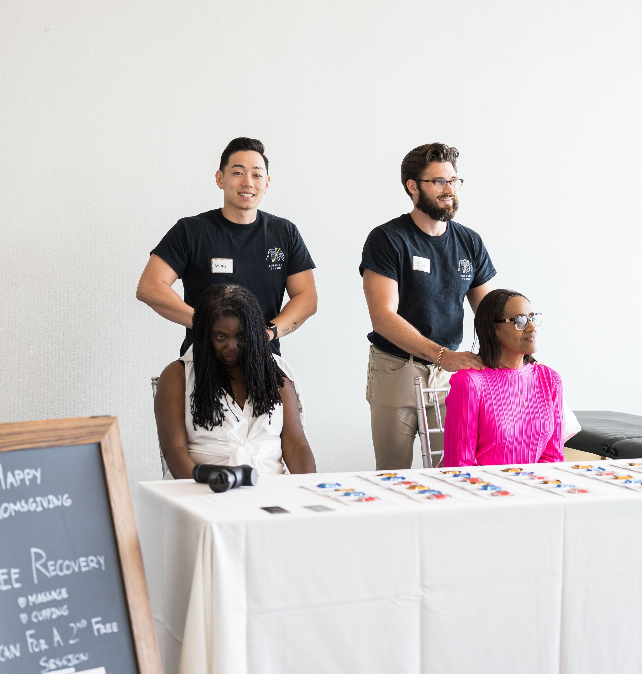 Two women receiving chair massages from massage therapists at a wellness event, with a table displaying informational materials and a chalkboard sign in the foreground.