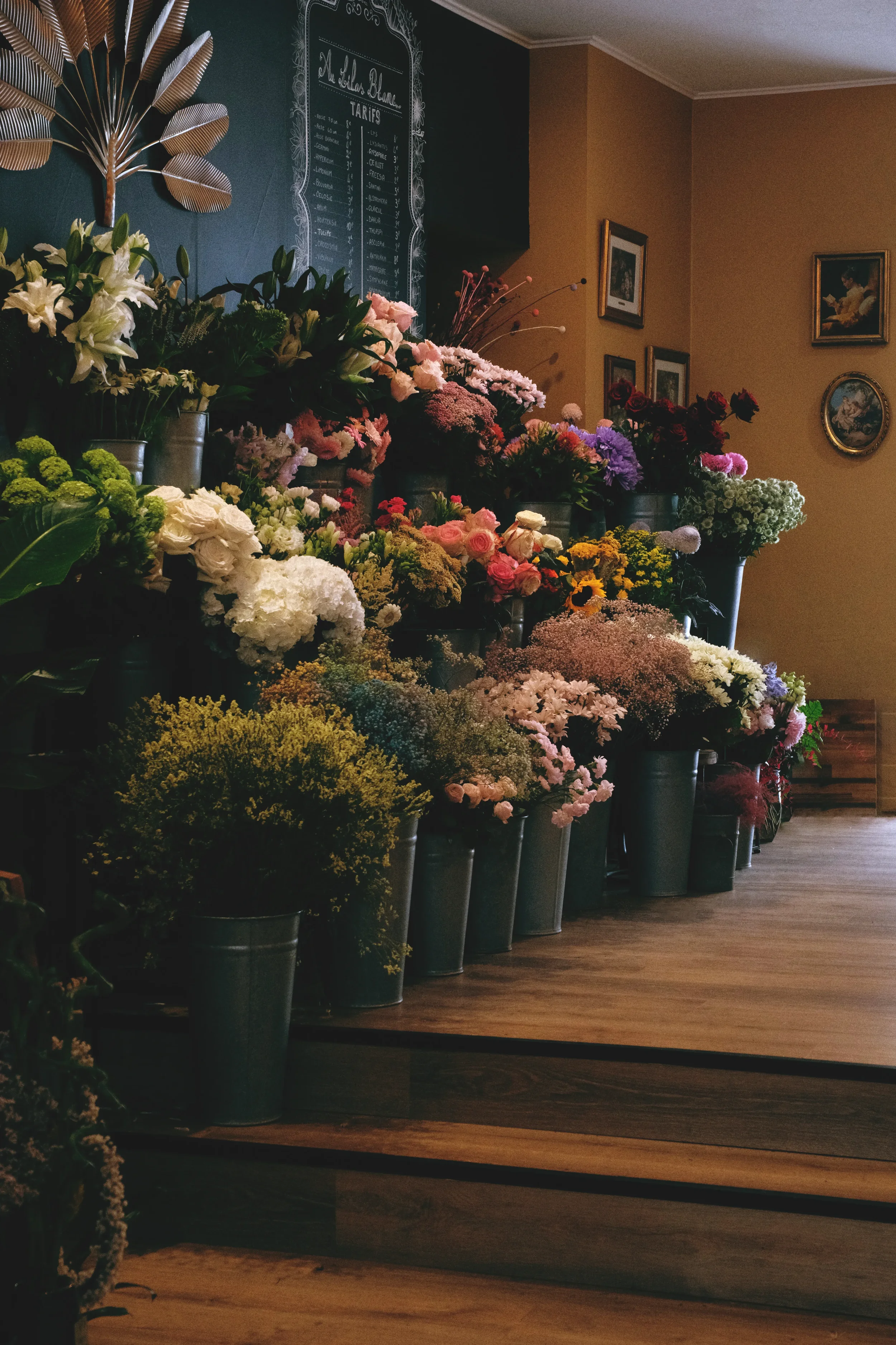 Une vitrine remplie de bouquets de fleurs colorées dans des vases en métal, avec un tableau noir au mur en arrière-plan et des cadres de peinture accrochés sur le mur.