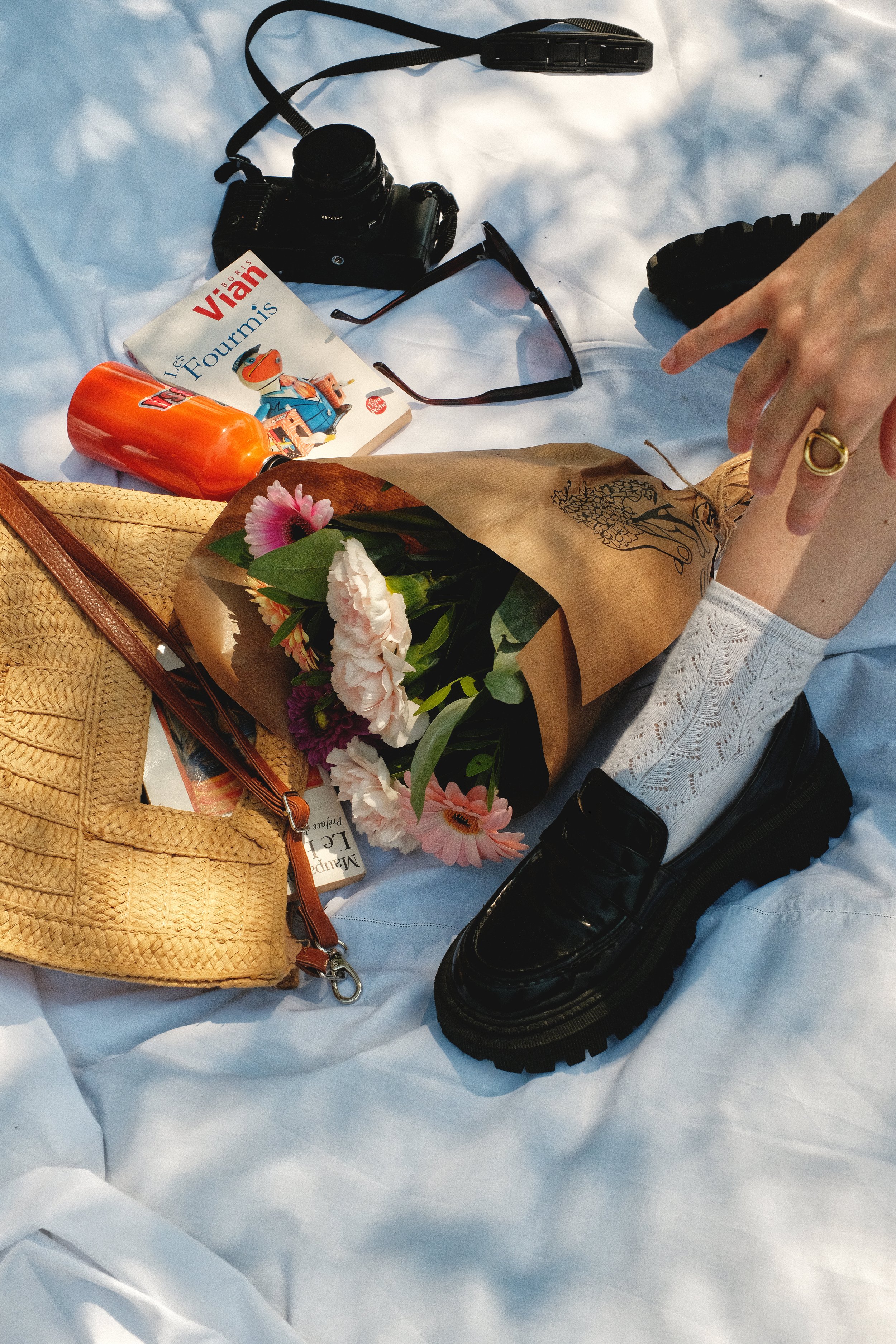 Une scène avec un bouquet de fleurs, un sac en paille, un livre , une bouteille orange, une paire de lunettes de soleil, un appareil photo, une chaussure noire, et une main qui ajuste une chaussette sur une personne. Editorial