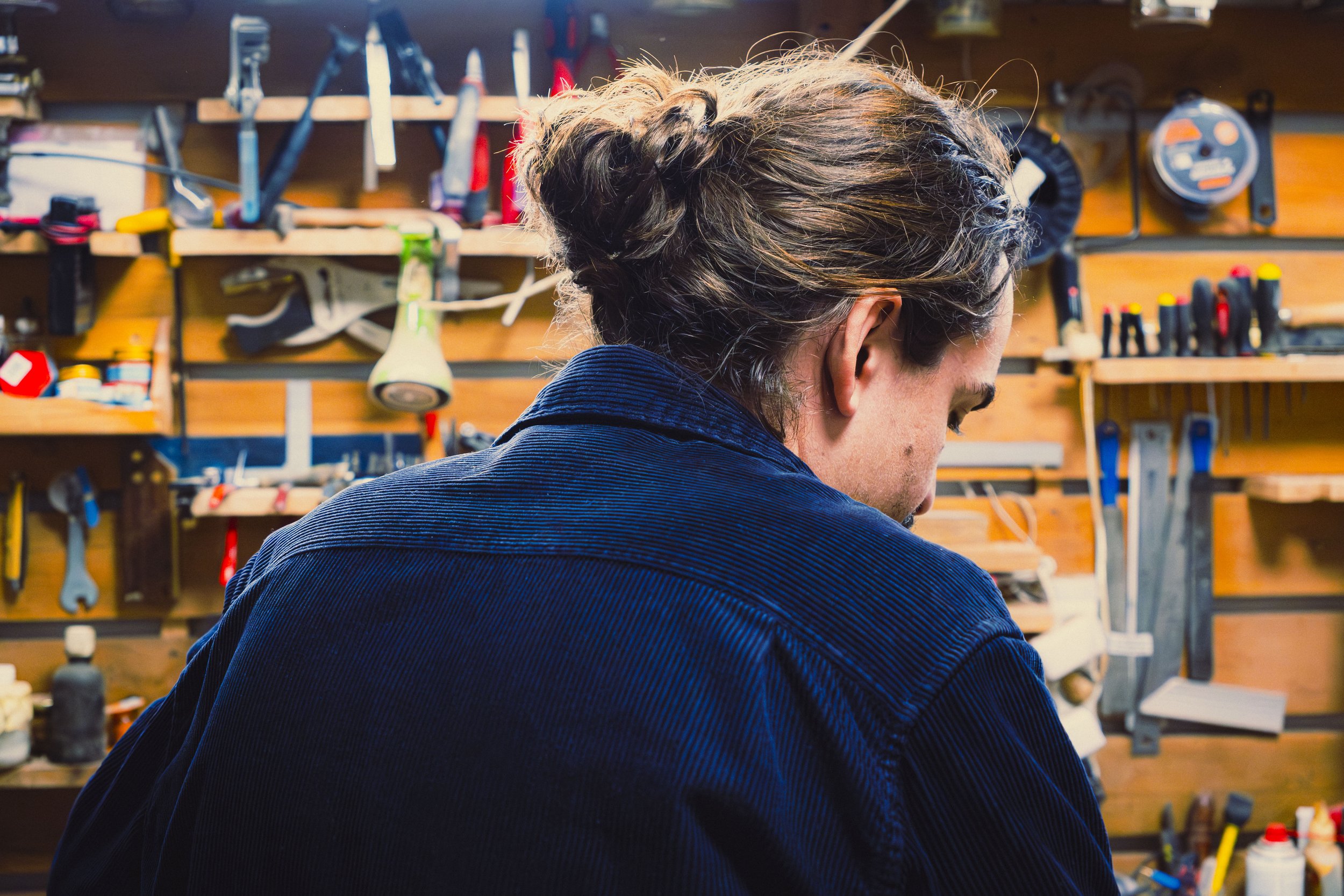Photo en couleur d'un homme de dos . Il est débout face à son atelier rempli d'outils.
