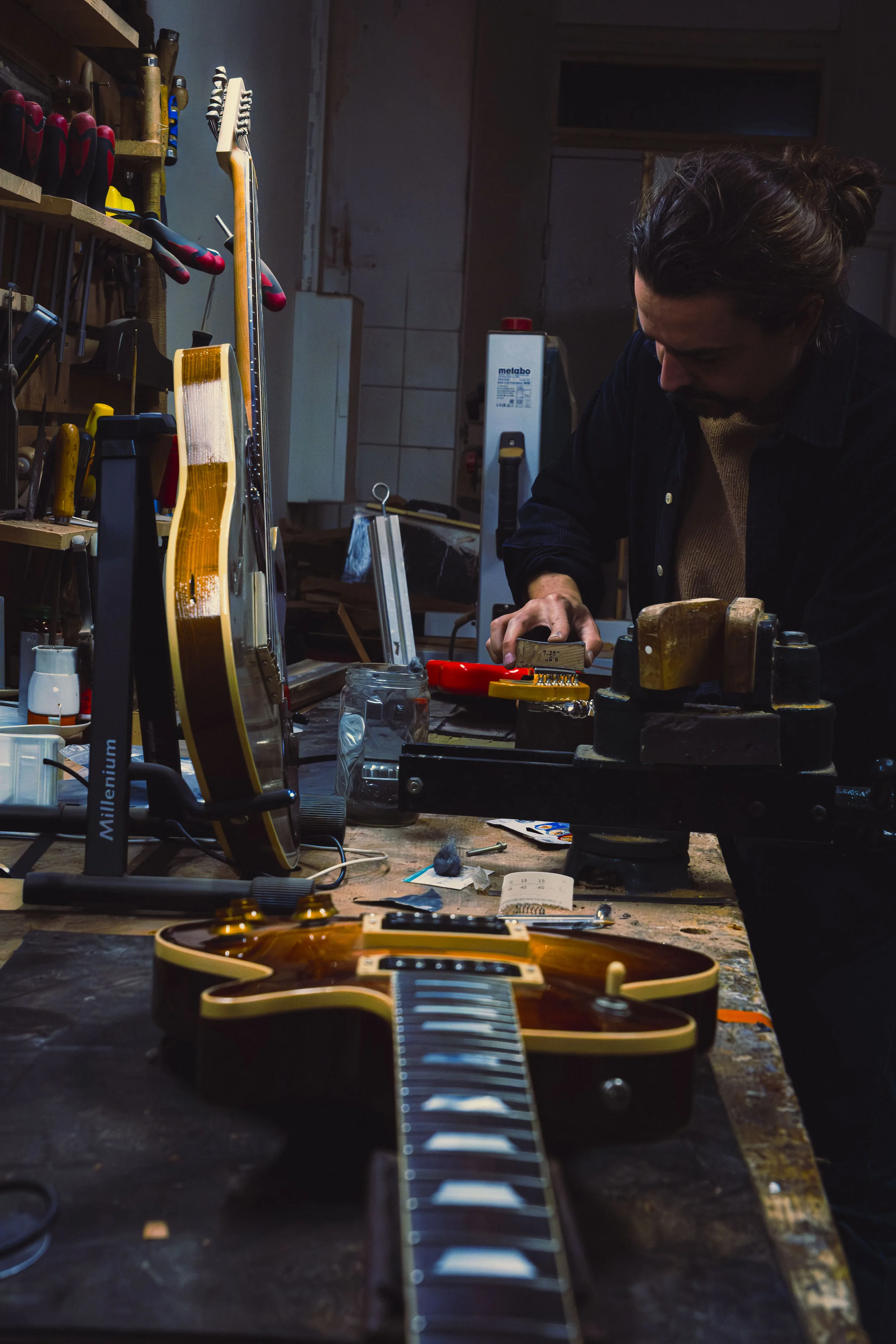 Un homme travaillant dans un atelier, en train de réparer une guitare électrique. La scène est visible depuis la tête de la guitare, avec divers outils et pièces autour.