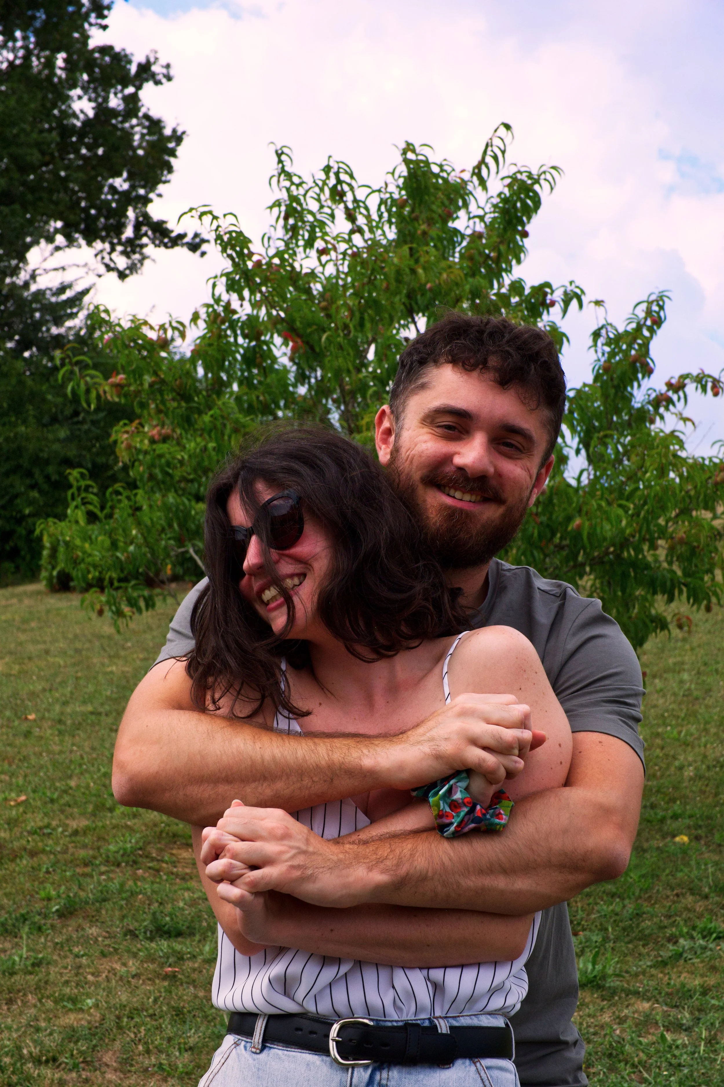 Un homme et une femme souriants s'enlacent amicalement dans un parc, avec un arbre en arrière-plan, sous un ciel partiellement nuageux. Portraits duo