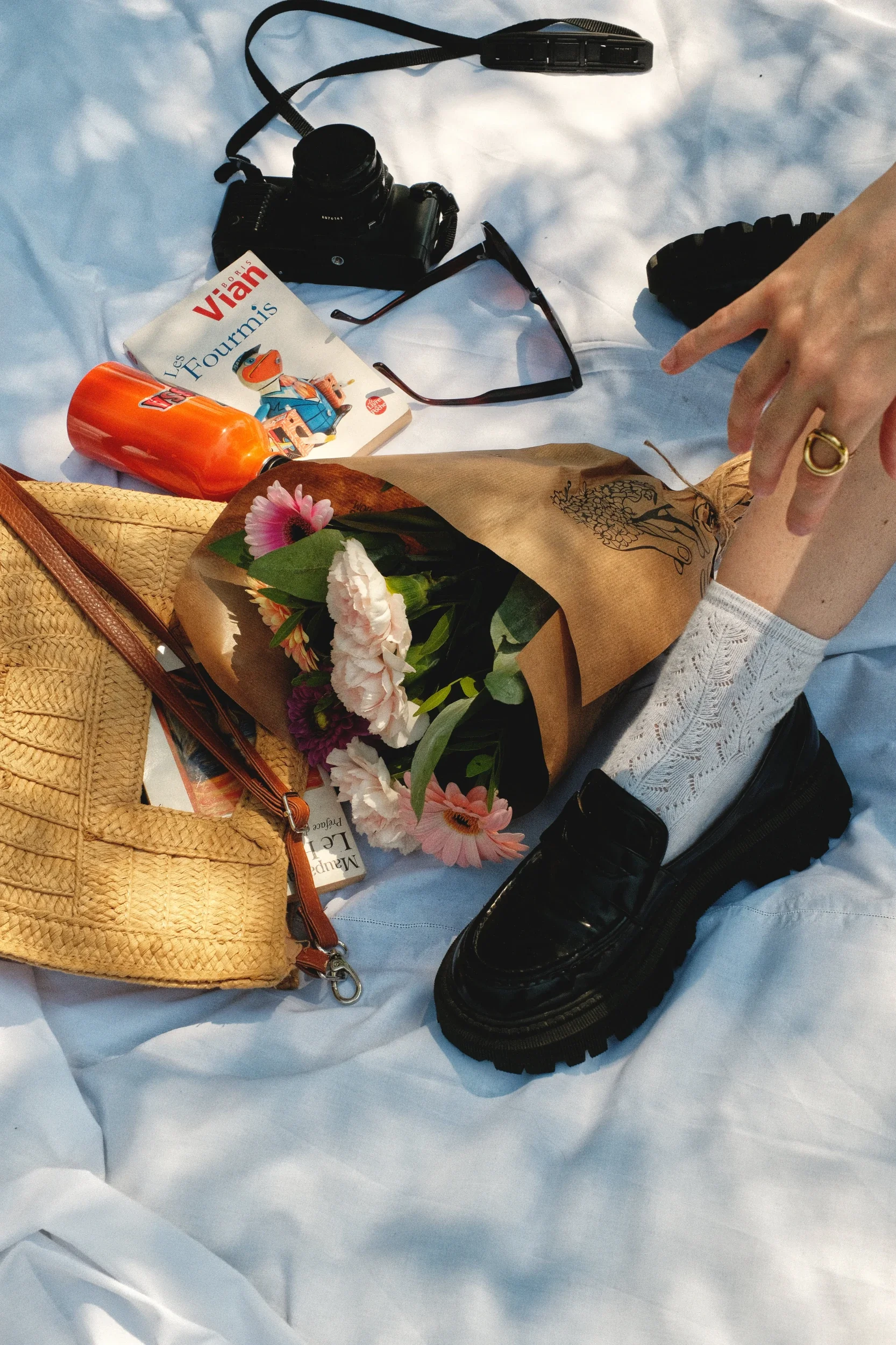 Une table blanche avec un appareil photo, des lunettes de soleil, un livre intitulé 'Les Fourmis' de Boris Vian, une gourde orange, un bouquet de fleurs et une personne portant une bague, une chaussure noire robuste.