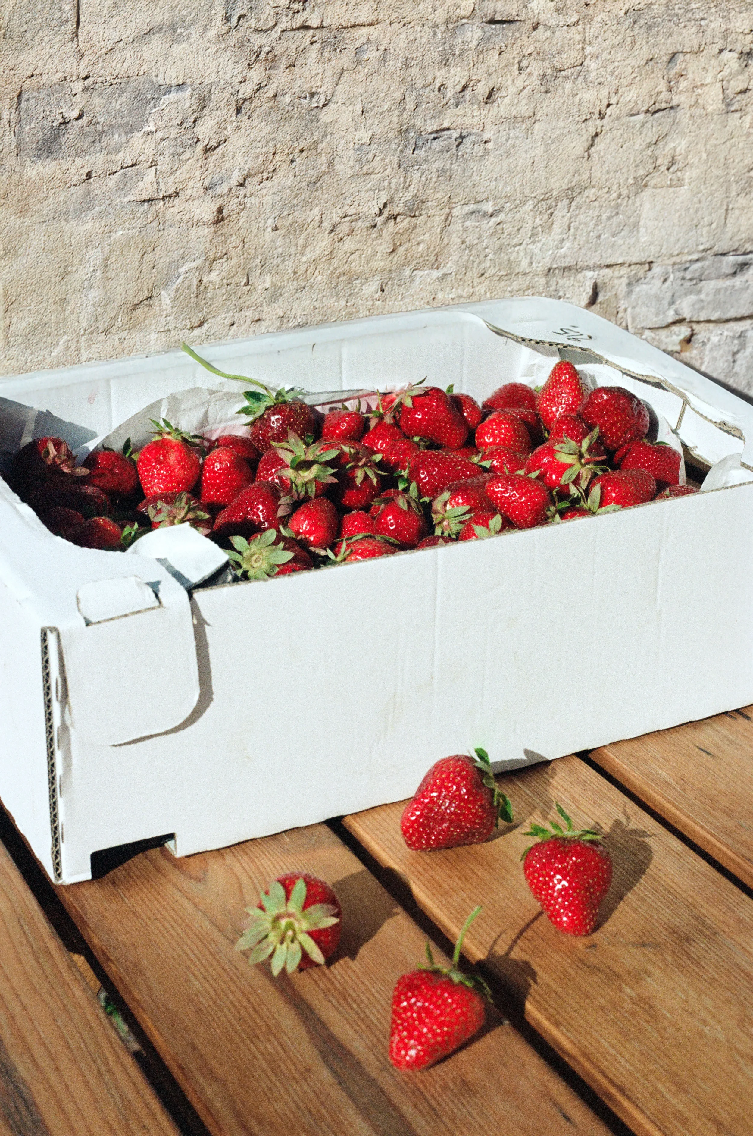 Une cagette carton blanc remplie de fraises rouges sur une table en bois. Quelques fraises sont dispersées sur la table devant la boîte. Reportage
