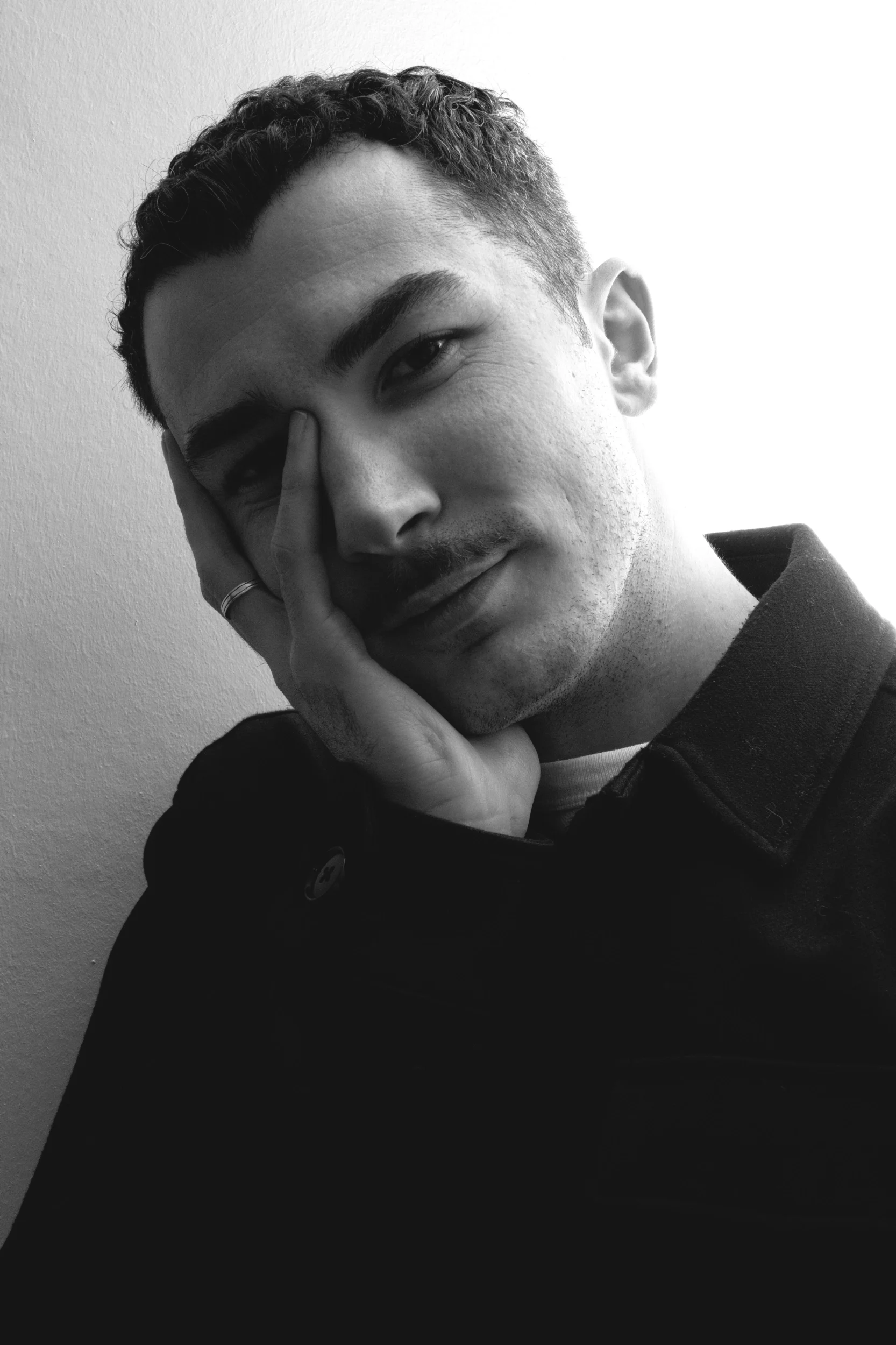 Un homme jeune à la peau claire, avec des cheveux courts et bouclés, portant un pull noir et posant sa tête dans sa main, regardant l'objectif dans une image en noir et blanc.