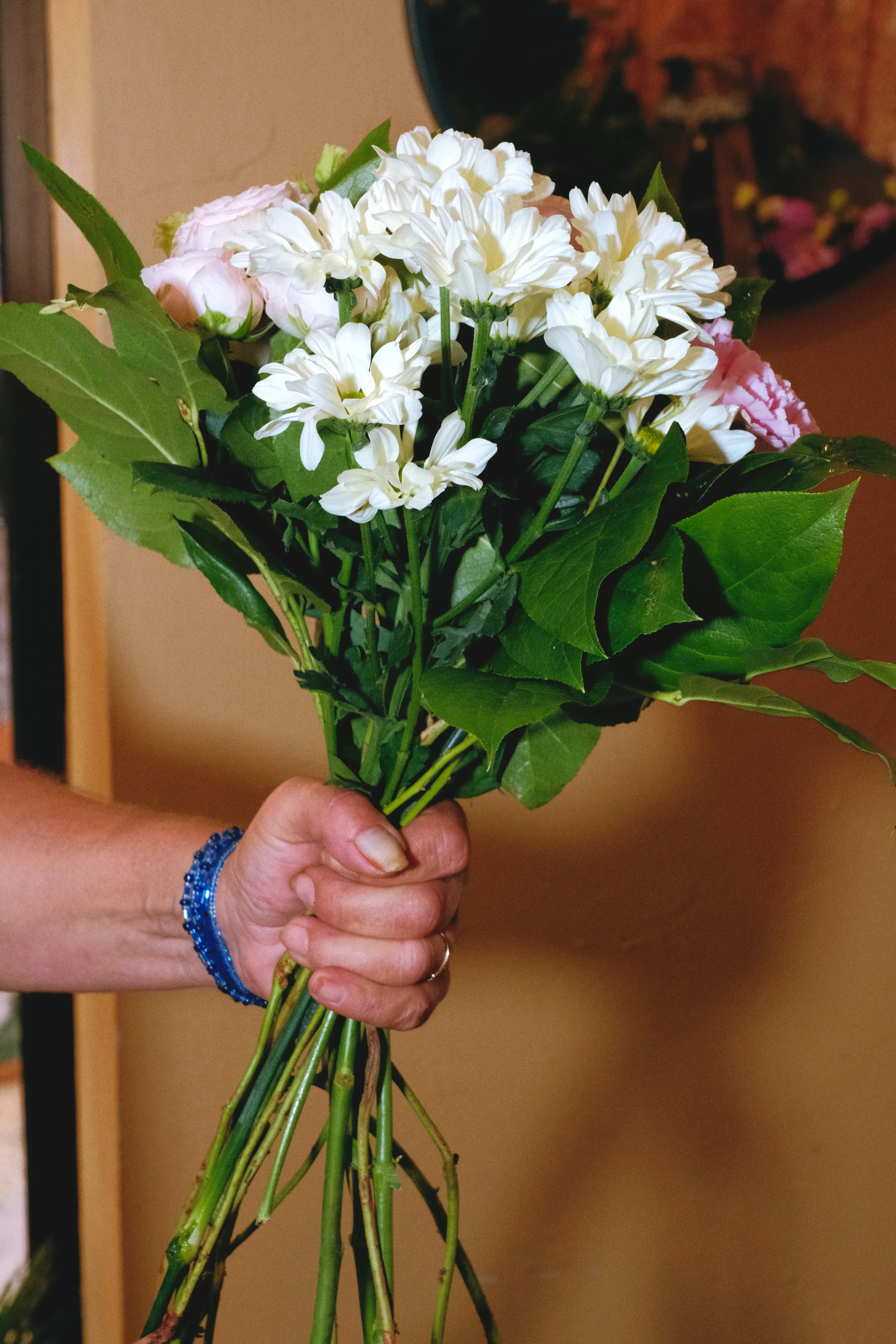 Main gauche, une main tenant un bouquet de fleurs comprenant des roses roses, des œillets blancs et des feuilles vertes. La personne porte un bracelet en perles bleues.