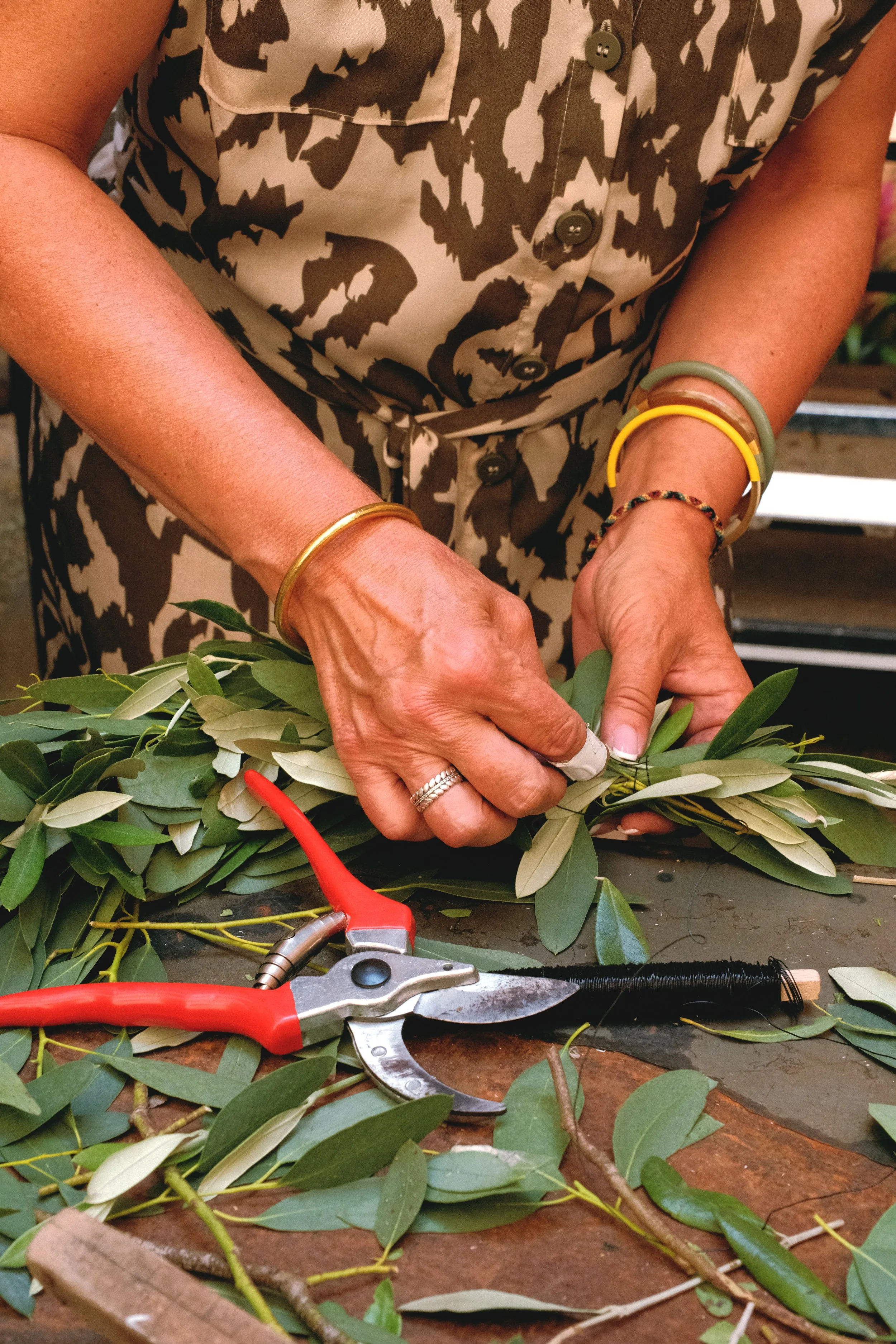 Une personne arrange des feuilles vertes avec des ciseaux de jardinage. La personne porte des bracelets colorés et une robe à motifs. Reportage