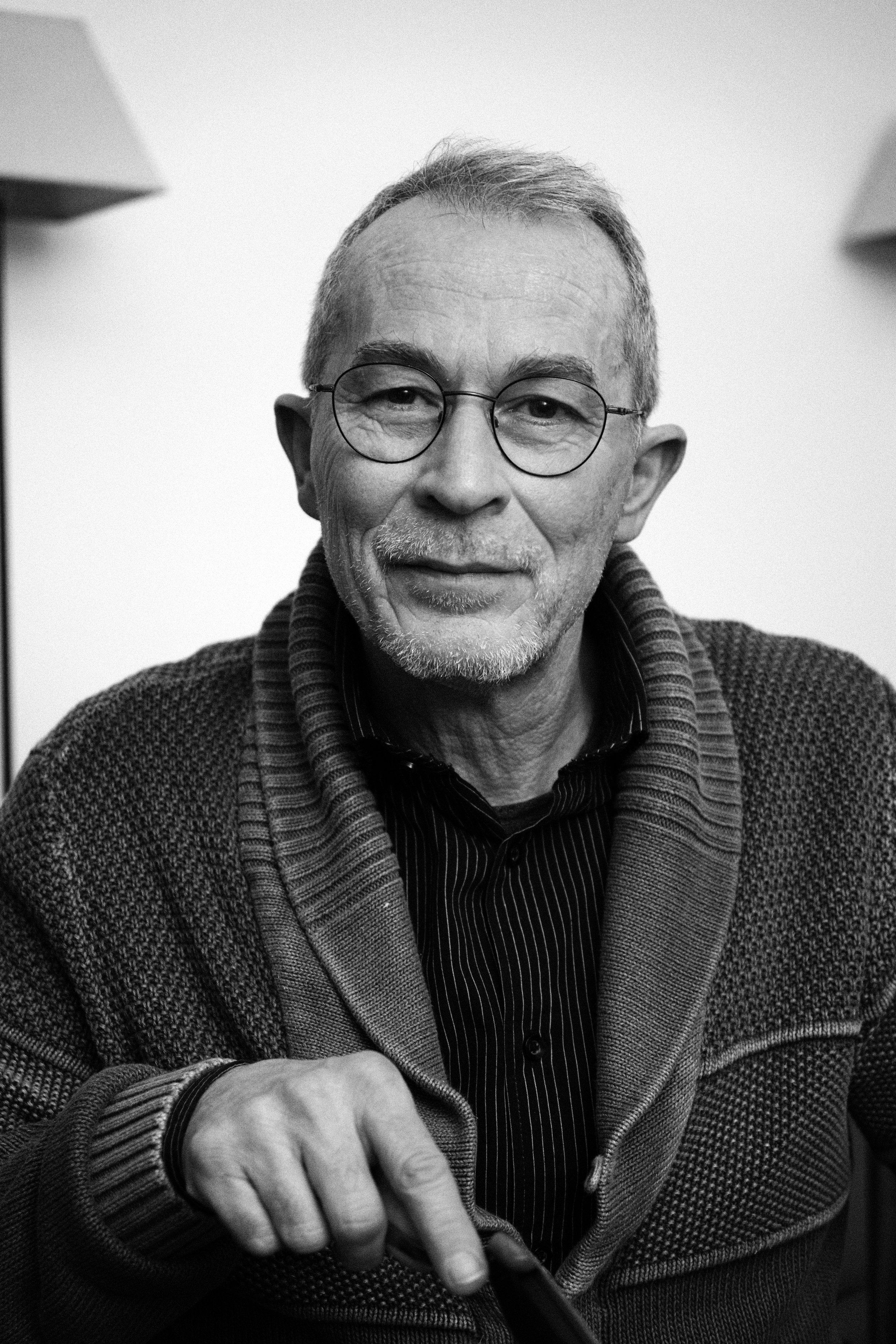 Portrait en noir et blanc d'un homme âgé, portant des lunettes rondes, un pull en laine et une chemise à rayures, souriant doucement. Portraits