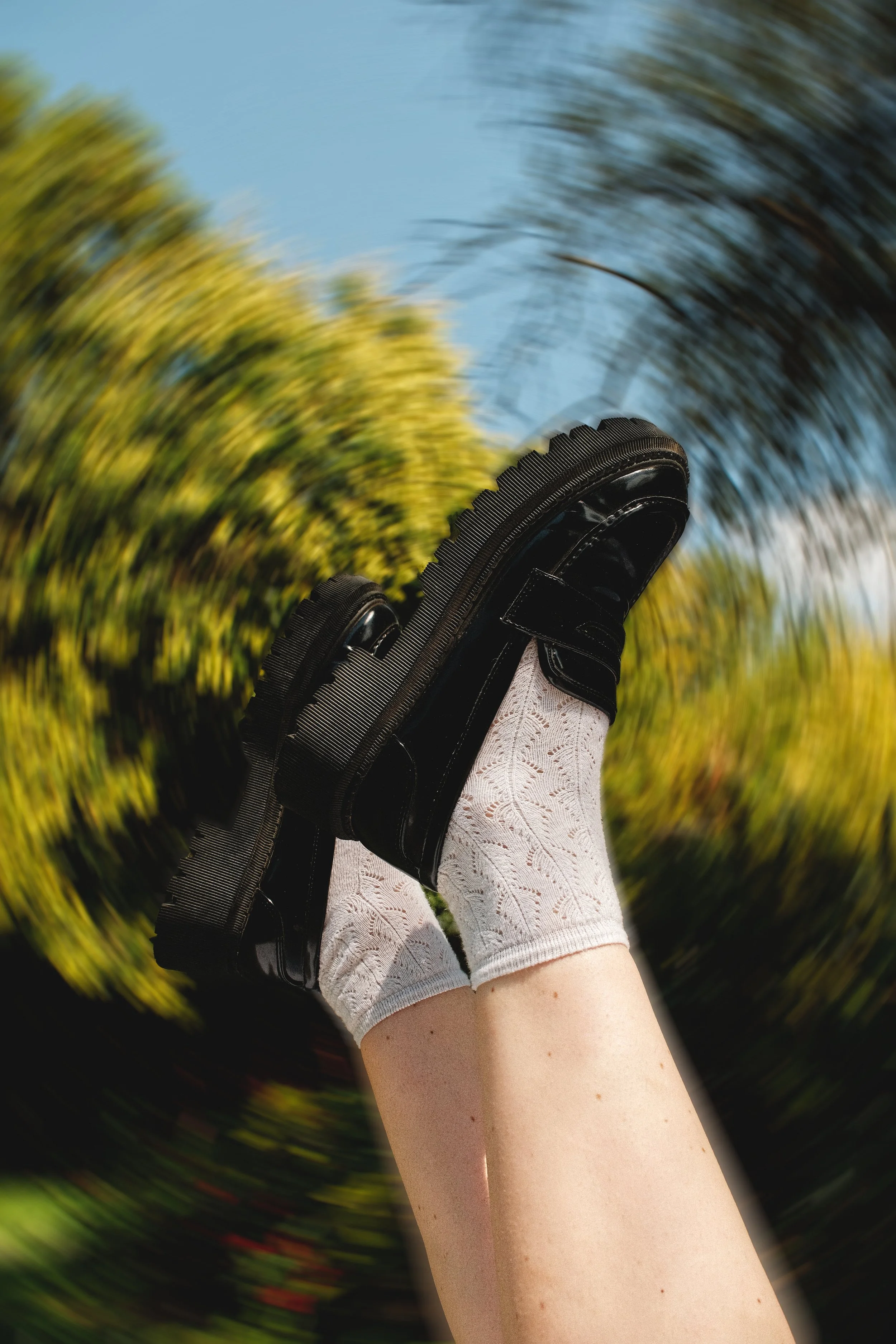 Une personne porte une chaussure noire avec des chaussettes blanches à motif, levée en l'air contre un ciel bleu et des arbres verts flous en arrière-plan. Editorial