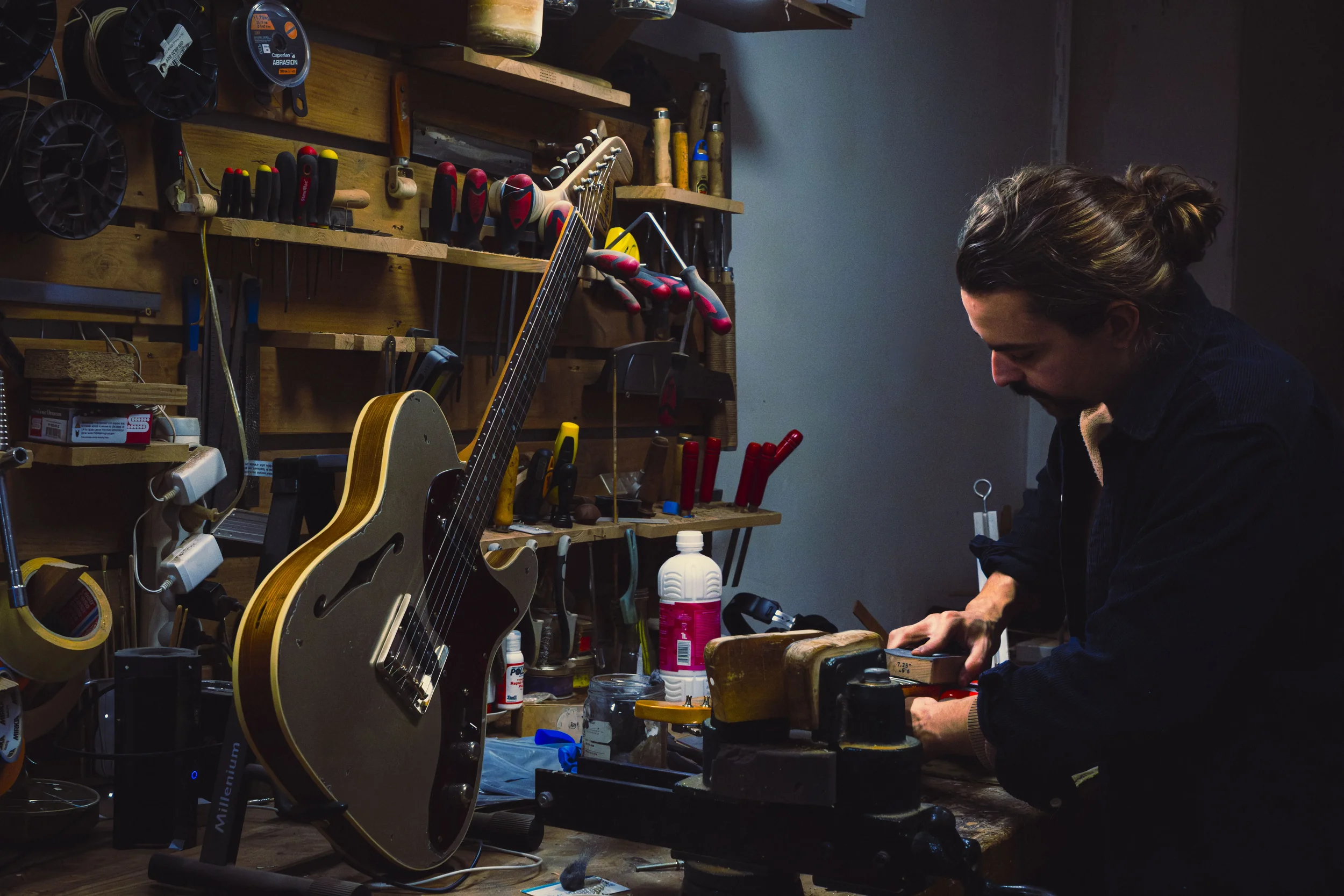 Photo en couleur d'un homme en train de vérifier raboter le manche d'une guitare. Il est débout au milieu de son atelier rempli d'outils. Reportage