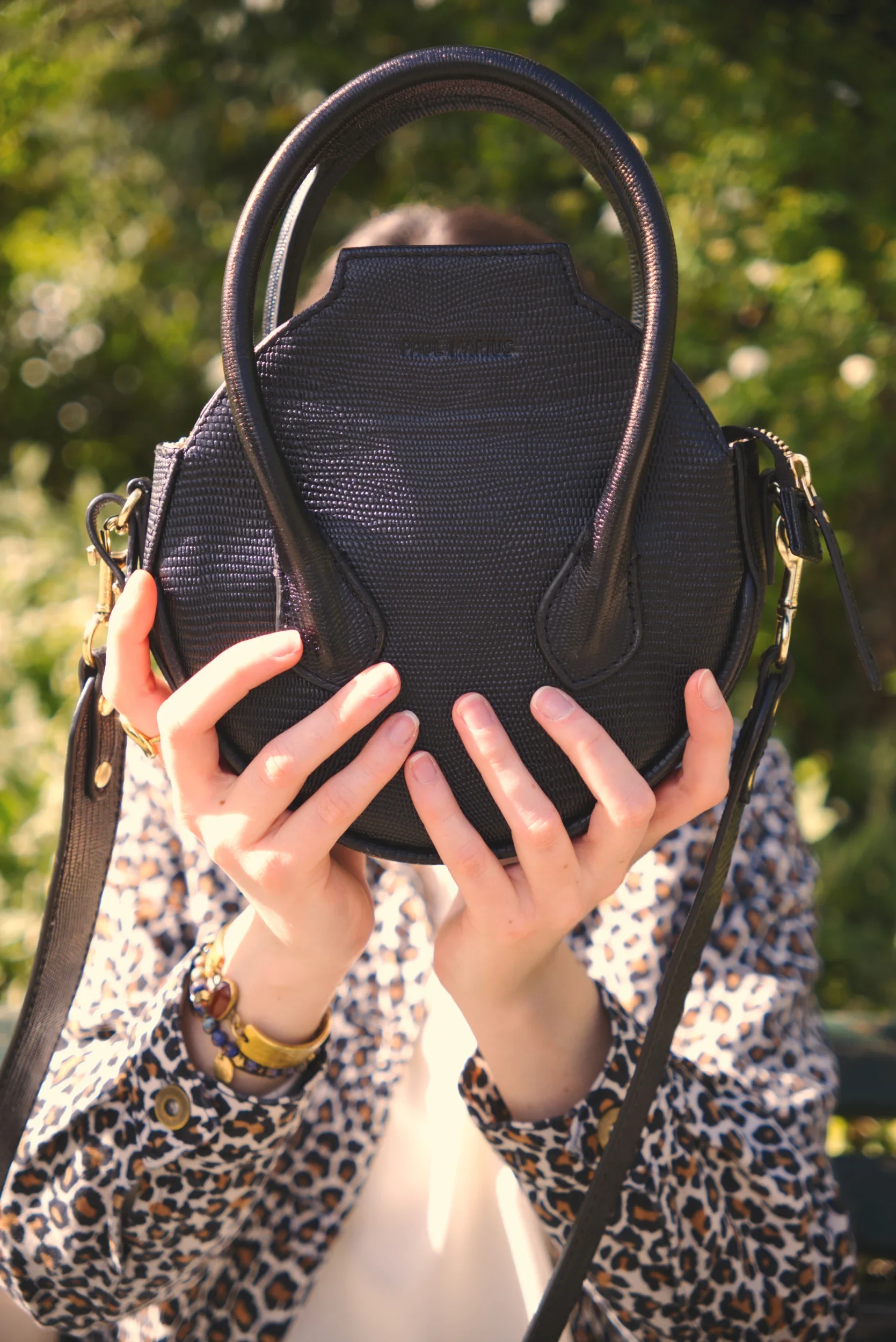 Une personne tient un sac à main noir en cuir avec des mains visibles, portant des bijoux, dehors avec un fond de verdure, portant une blouse à motifs léopard.