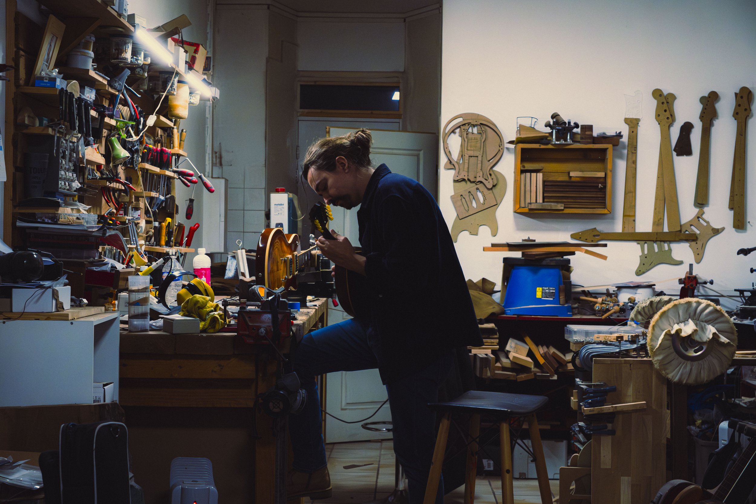 Photo en couleur d'un homme tenant une guitare et en train de vérifier qu'elle est accordée. Il est débout au milieu de son atelier rempli d'outils, de manches de guitare. Reportage