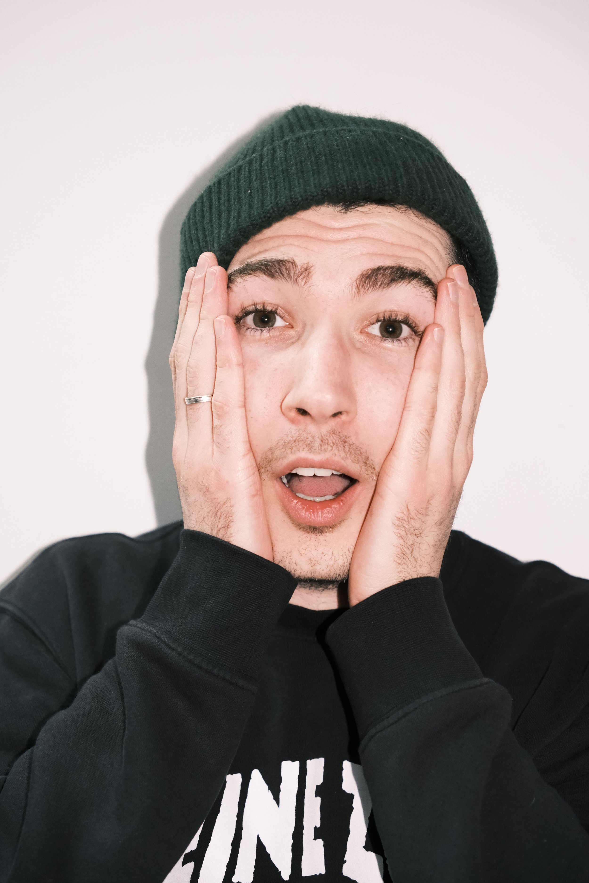 Jeune homme surpris avec une main sur chaque côté du visage, portant un bonnet vert et un sweat noir, avec un fond blanc.