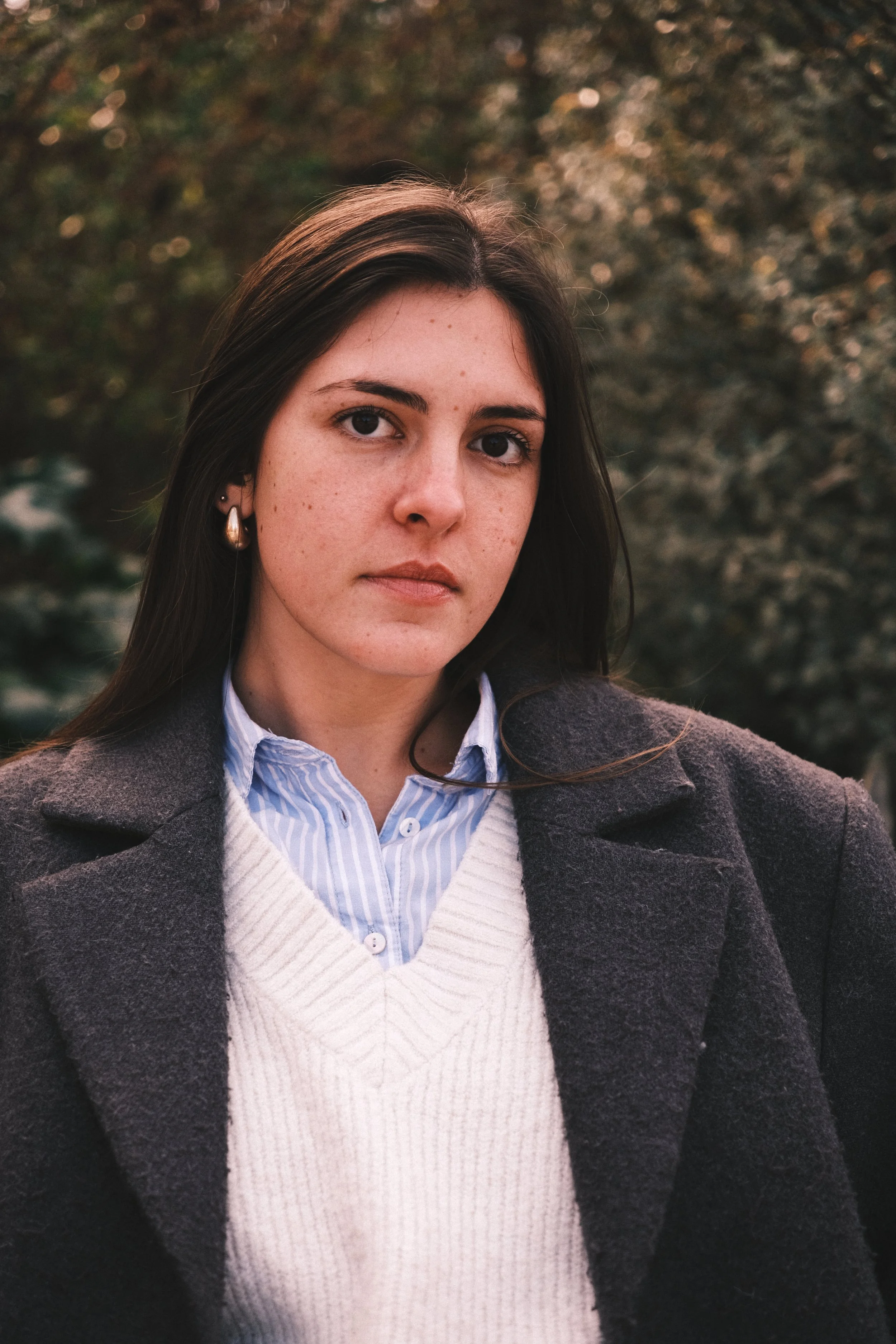 Jeune femme avec des vêtements d'automne, foncée, avec des cheveux longs et bruns, portant des boucles d'oreilles, posant à l'extérieur avec un arrière-plan de feuilles d'arbres. Portraits