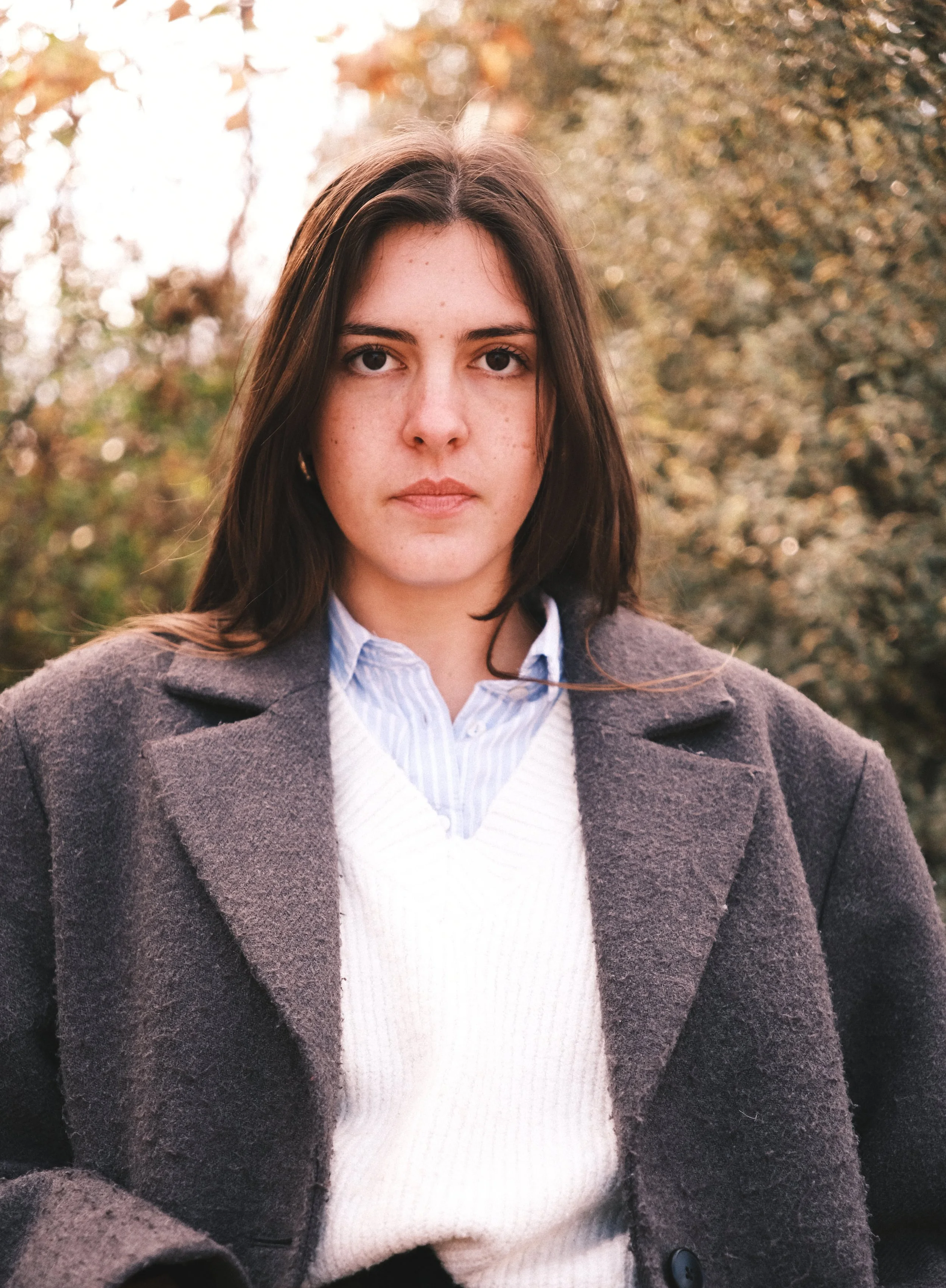 Jeune femme aux cheveux bruns, portant une veste grise et un pull blanc, debout dans un paysage automnal avec arbres aux couleurs d'automne. Portraits