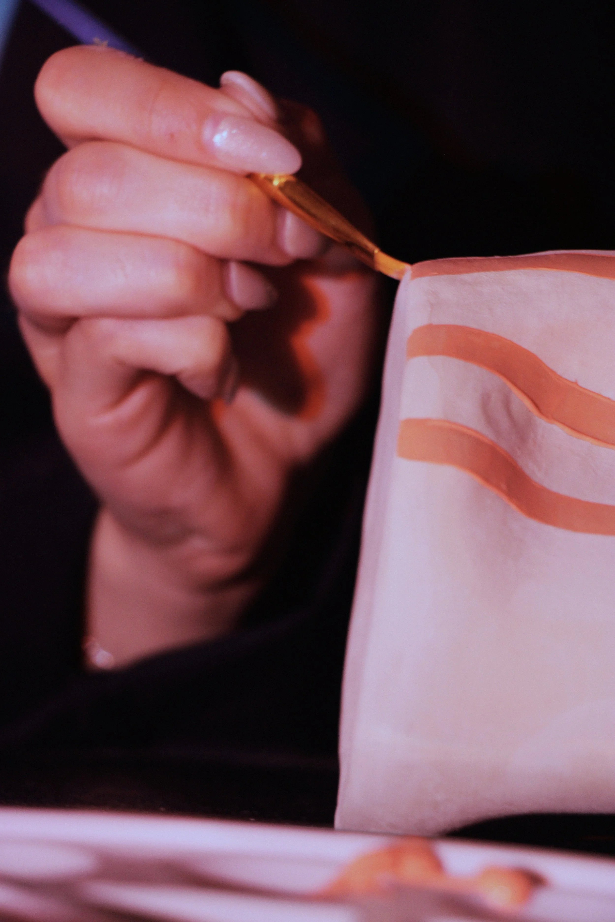 Une main tenant un pinceau et peignant des bandes sur un objet en papier ou tissu, couleurs rosées et orangées. Reportage