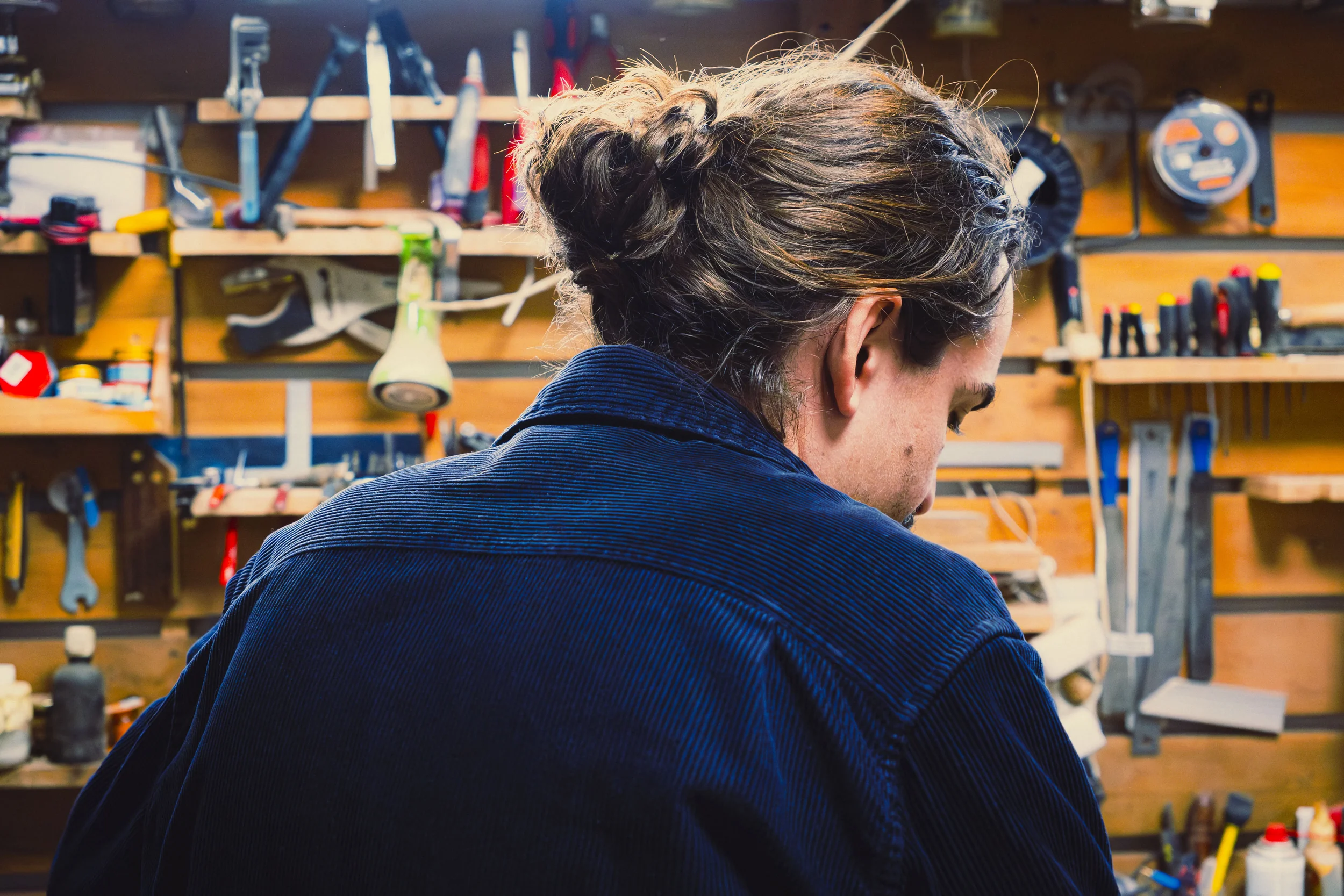 Photo en couleur d'un homme de dos . Il est débout face à son atelier rempli d'outils.