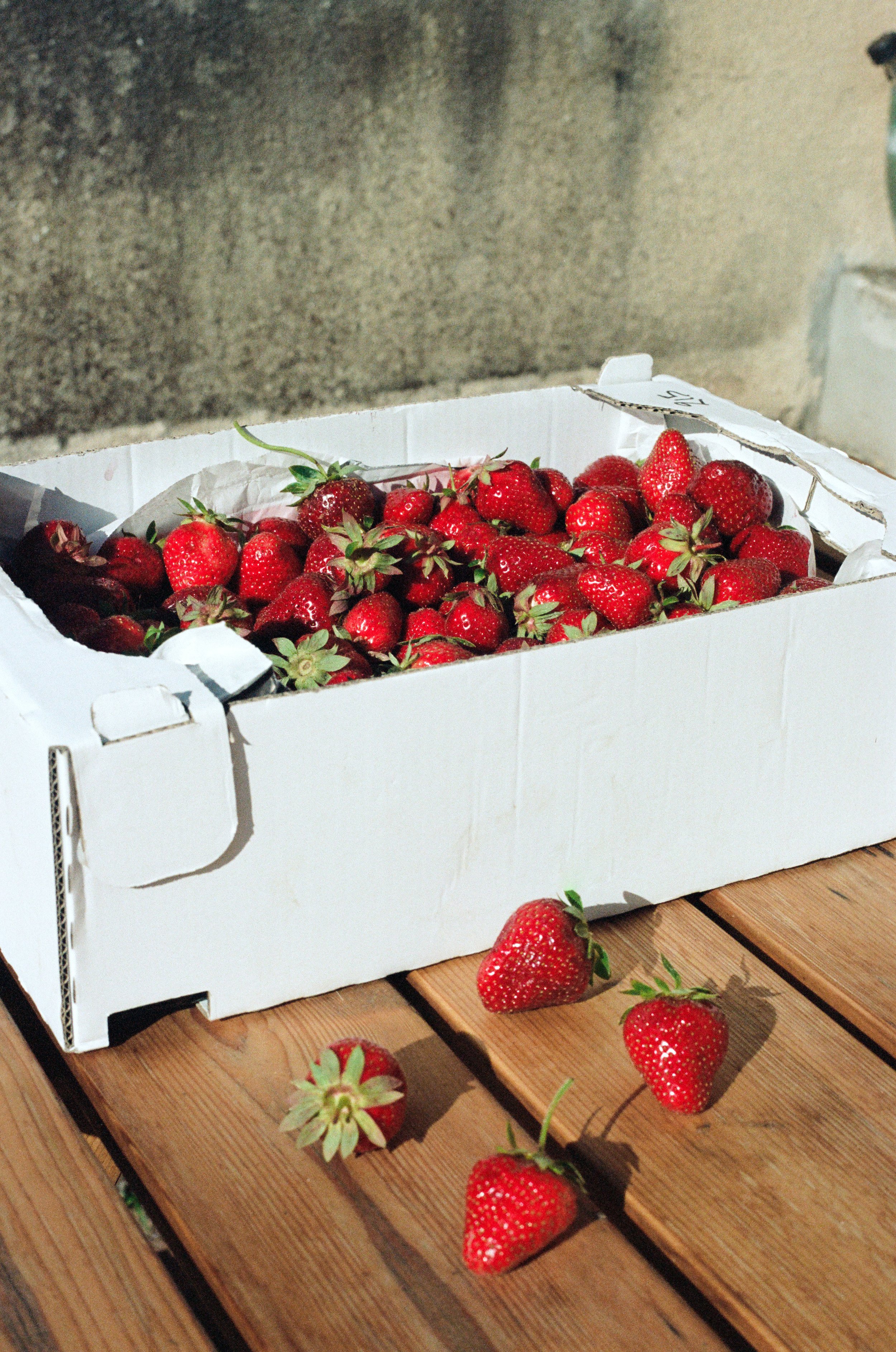 Une boîte en carton blanc remplie de fraises rouges sur une table en bois. Quelques fraises sont dispersées sur la table devant la boîte. Reportage