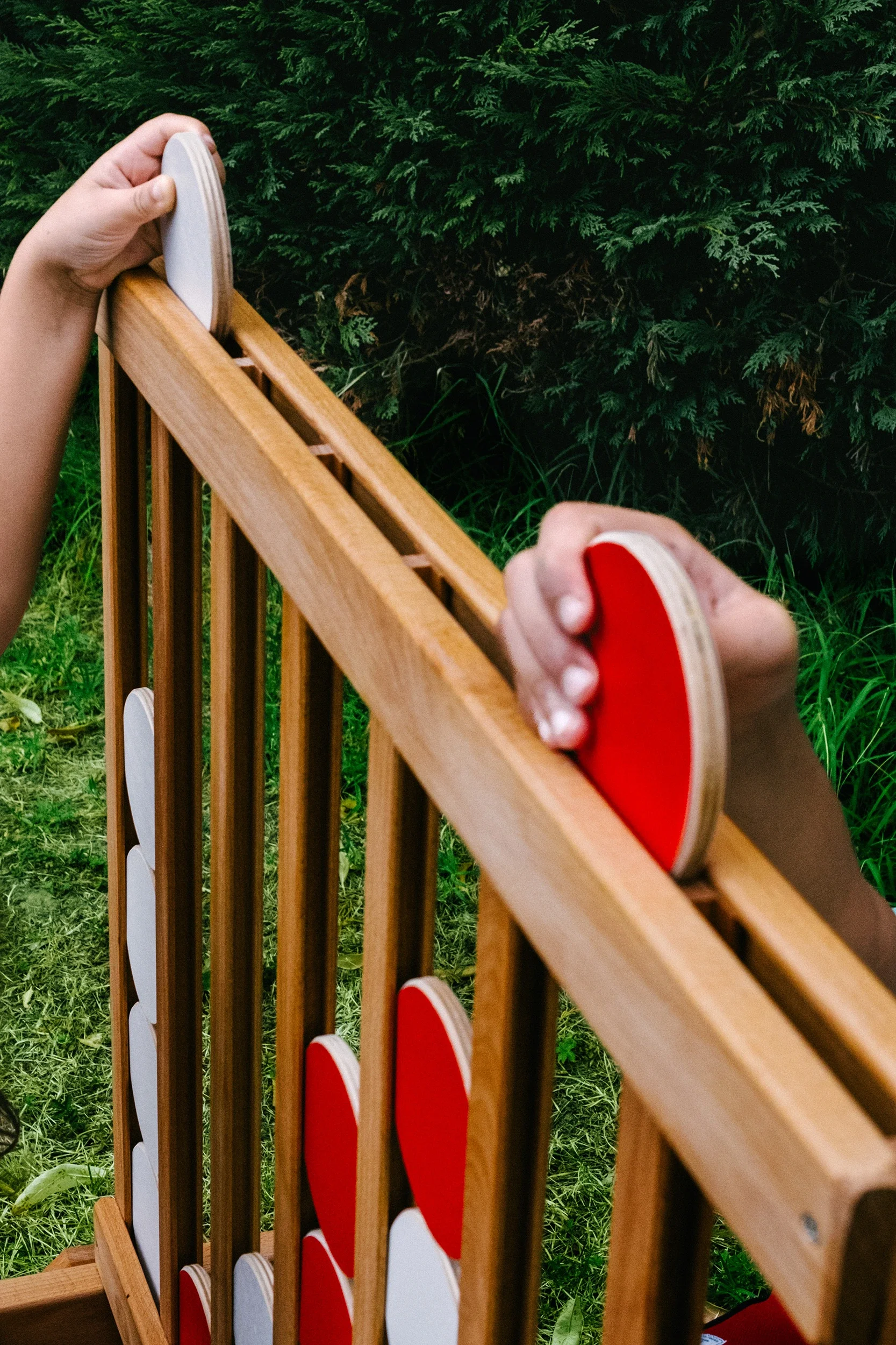 Deux mains tiennent des palets en bois, ublanc rouge, posés sur une structure en bois, avec une haie verte en fond.