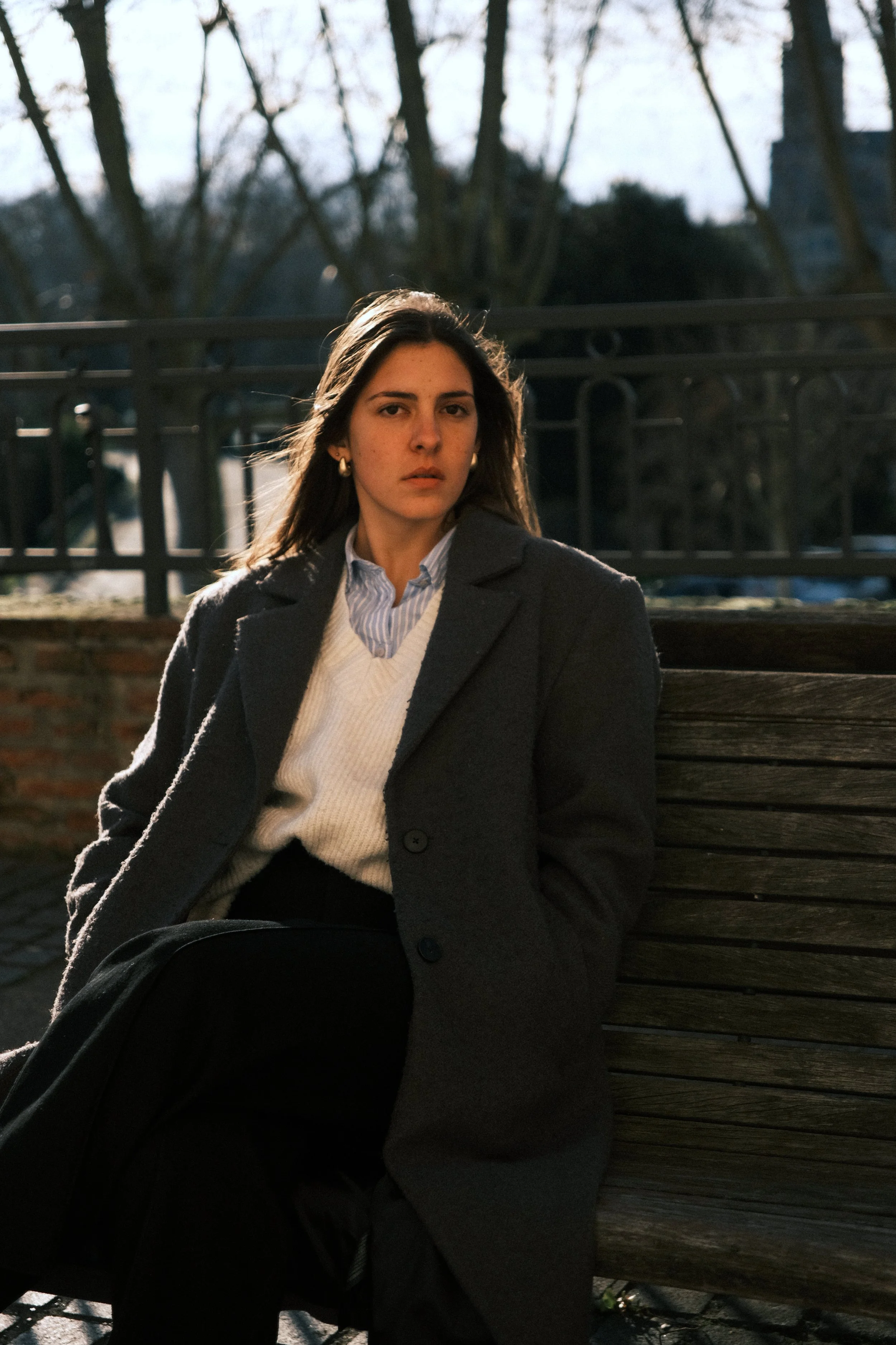 Femme assise sur un banc en bois dans un parc pendant la journée, portant un manteau gris, un pull blanc, et une chemise bleue rayée, avec des arbres nus en arrière-plan. Portraits