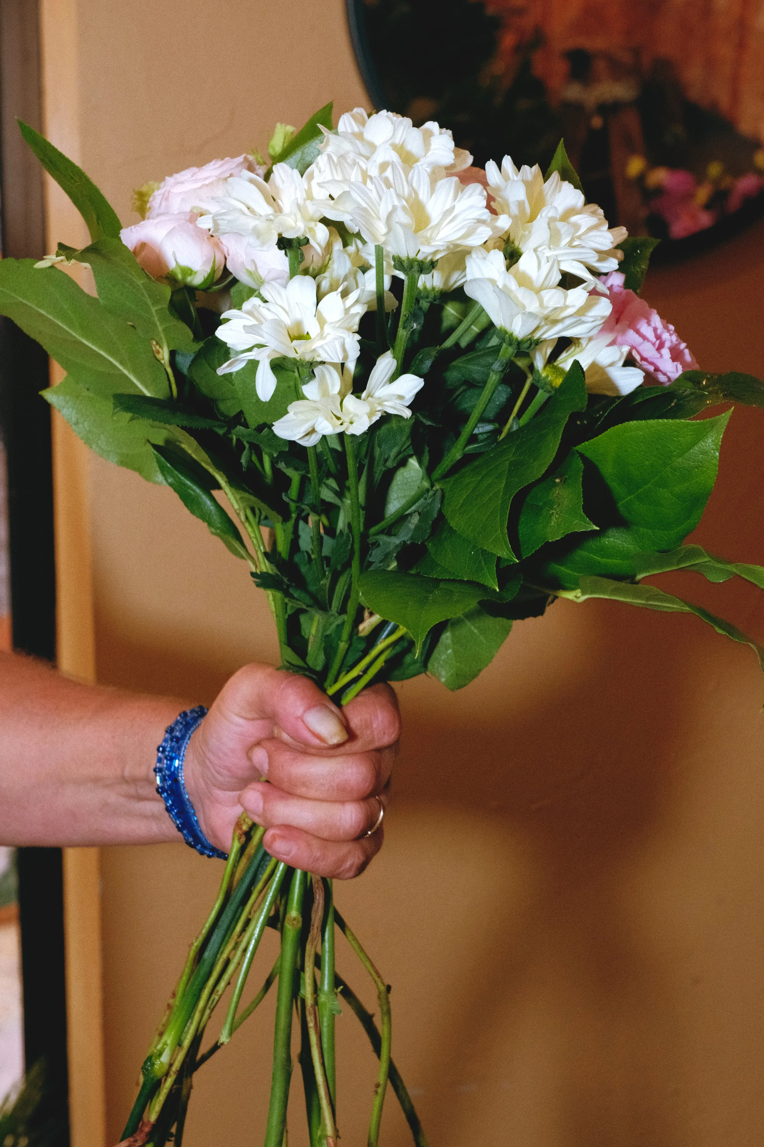 Main tenant un bouquet de fleurs blanches et roses, avec de grandes feuilles vertes, tenu par une personne portant un bracelet en perles bleues. Reportage